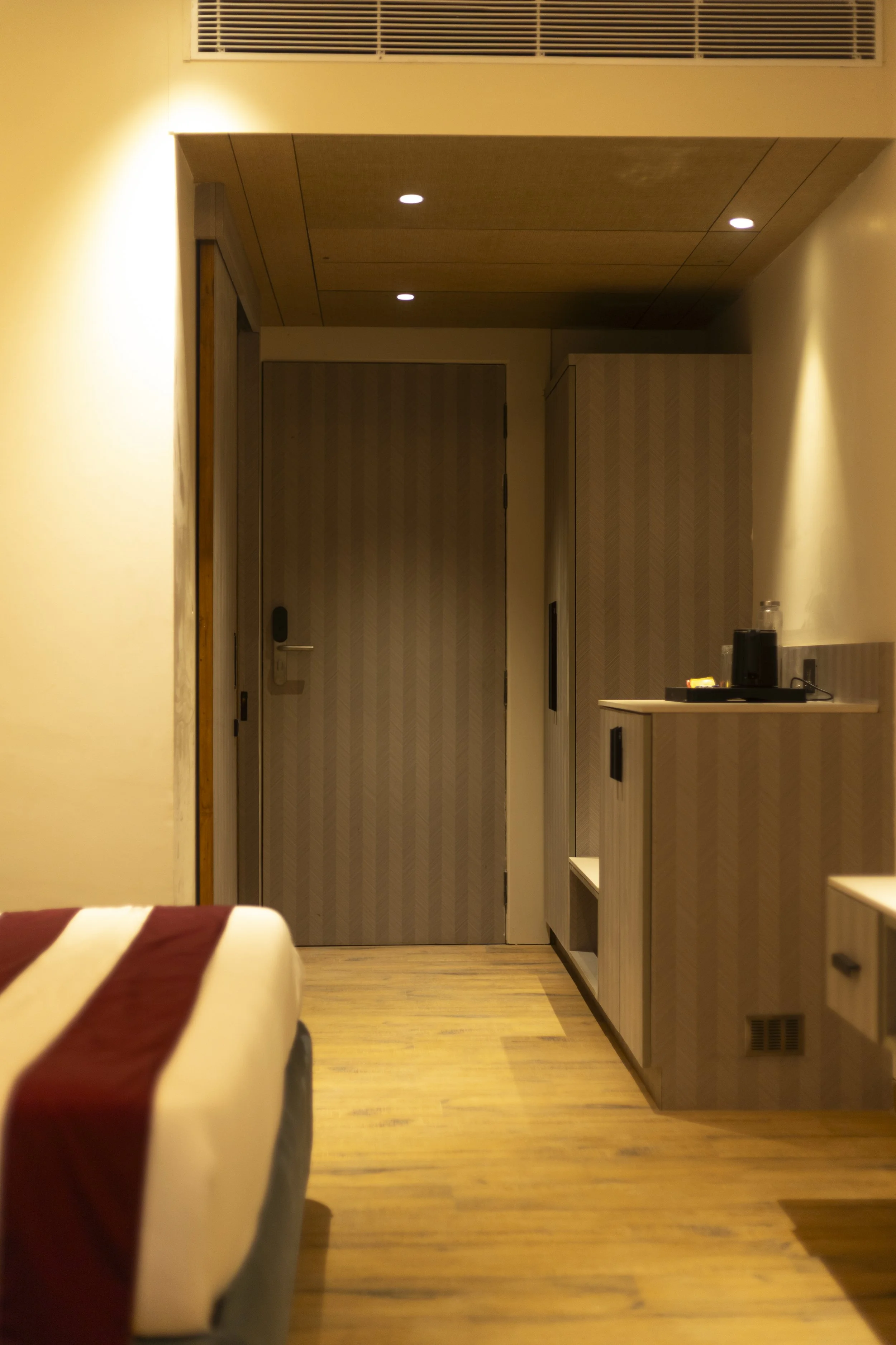 Empty hotel room with closed door, wooden ceiling panels, and a small shelf holding a coffee maker, cups, and bottles.