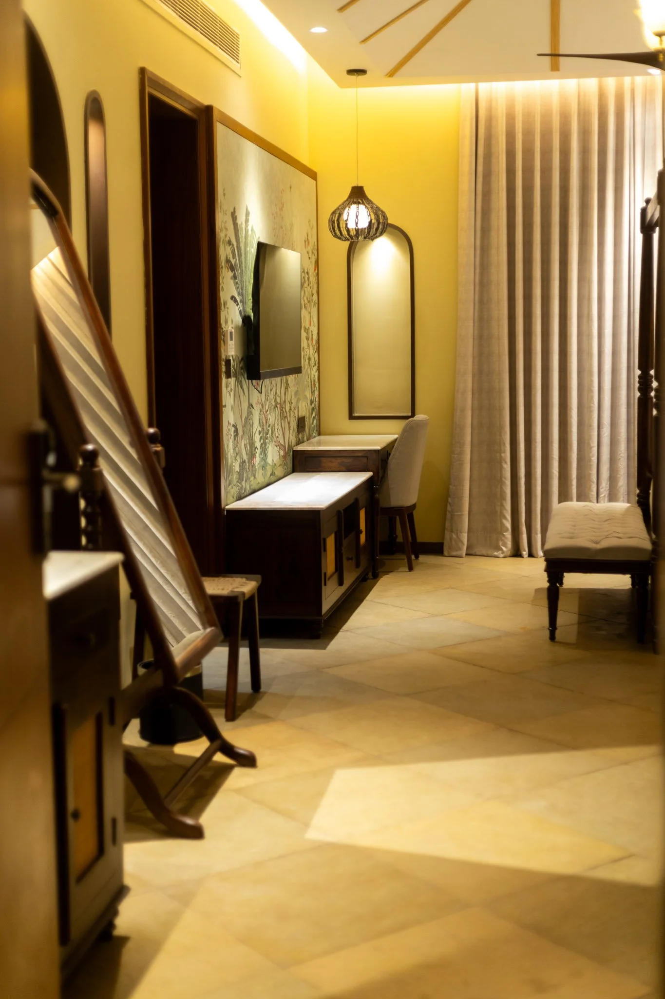 Interior of a hotel room with yellow walls, a flat-screen TV mounted on the wall, a small desk and chair, a mirror on the wall, and curtains covering a window or balcony door.