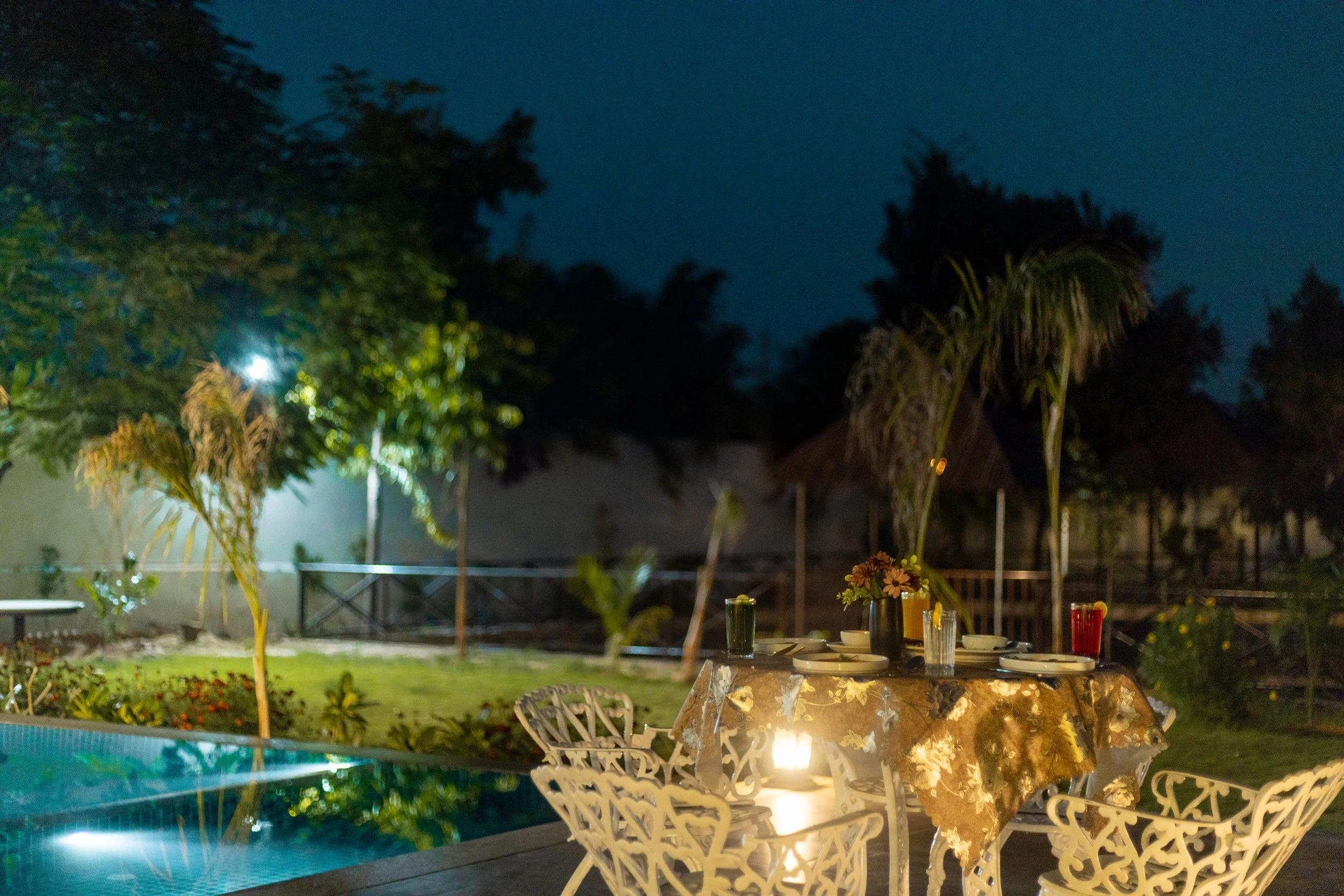 An outdoor dinner setup at night with a round table covered with a floral tablecloth, set with plates, glasses, and a small flower vase, surrounded by white ornate chairs, next to a swimming pool with lights, and a garden with trees and bushes.