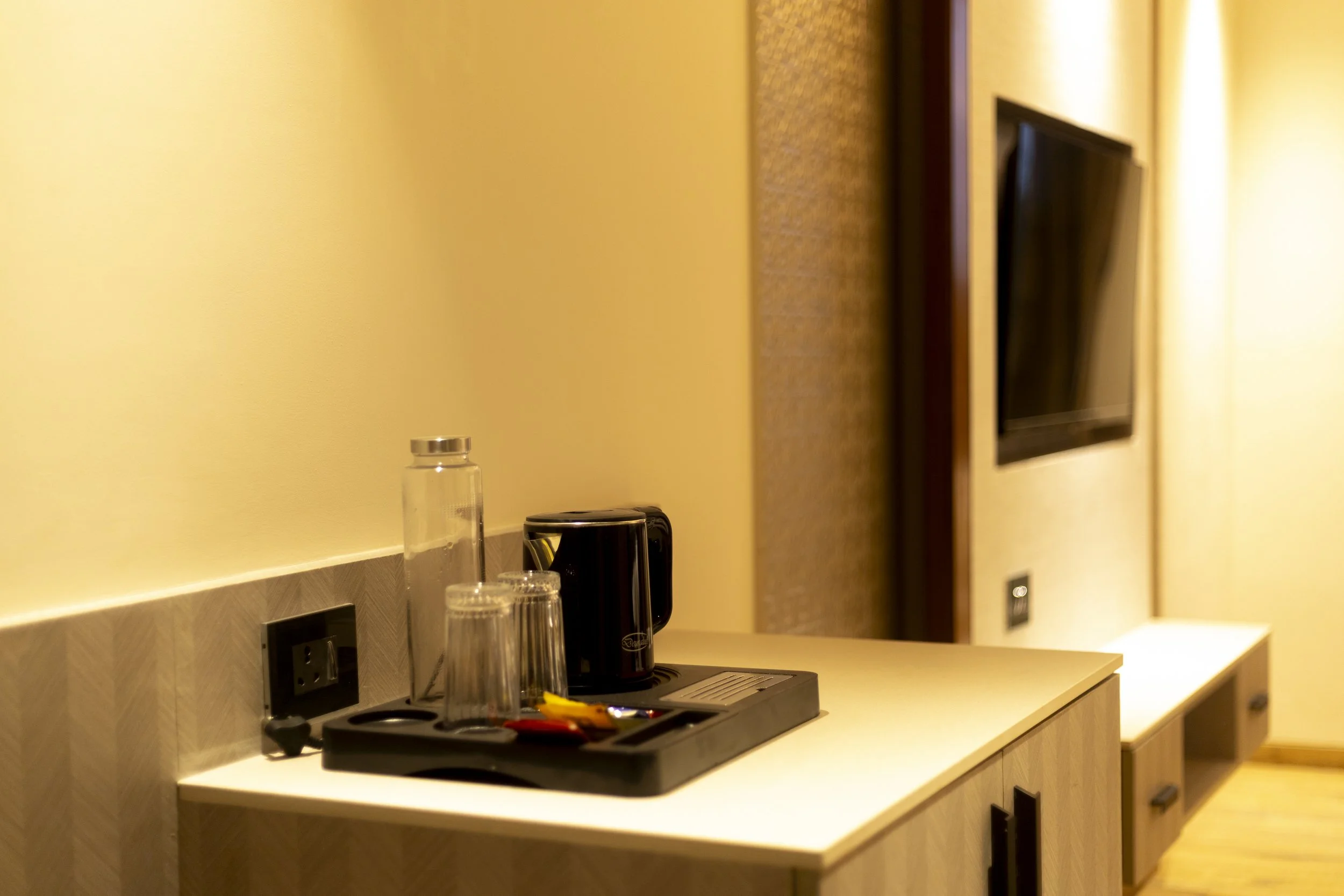 Coffee station on a wooden countertop with a glass water bottle, glasses, and a black electric kettle, in a hotel room with a wall-mounted TV and beige walls.