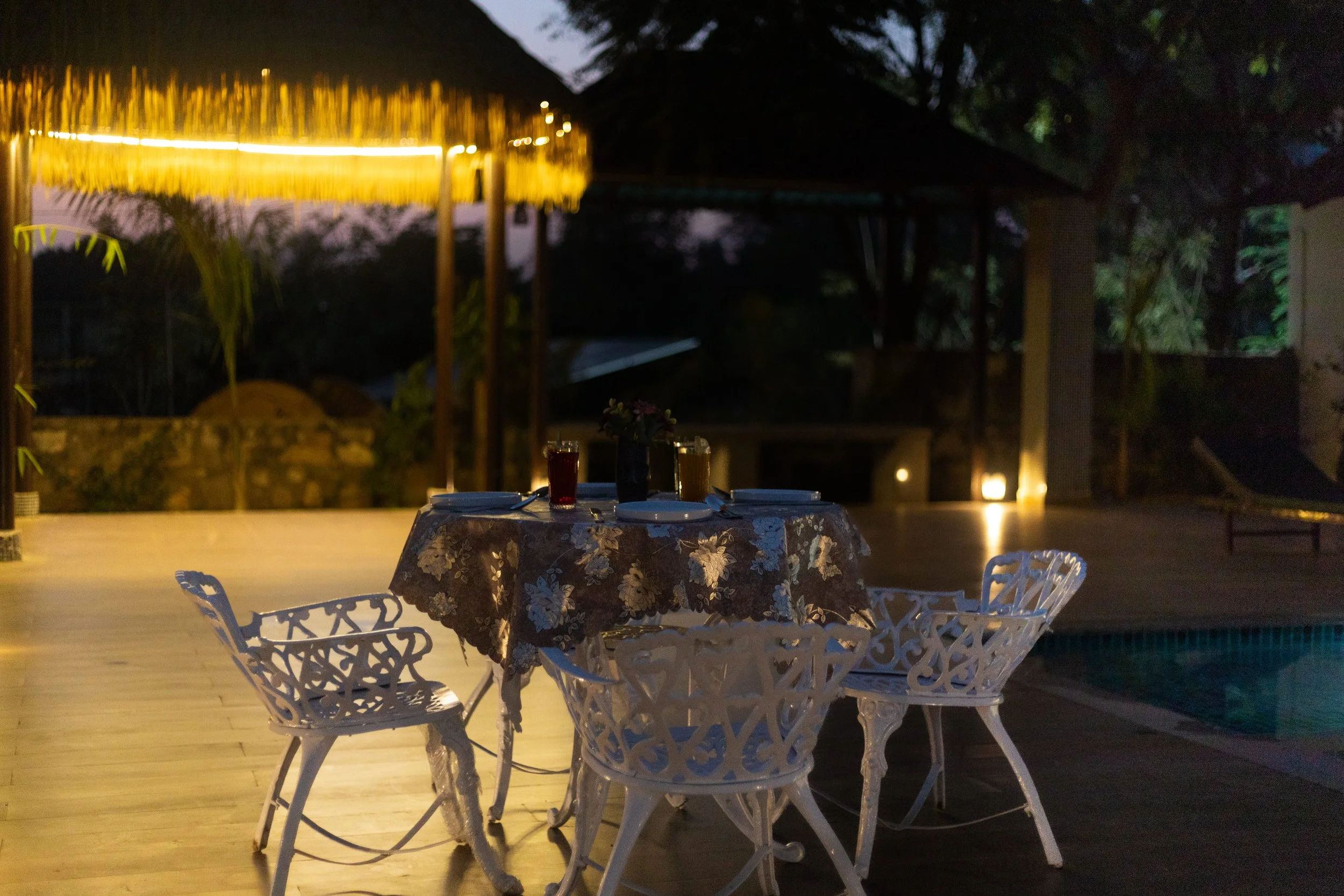 Outdoor dining table set with four white chairs, tablecloth, drinks, and flowers, in a backyard with a pool and tiki hut, at night with ambient lighting.