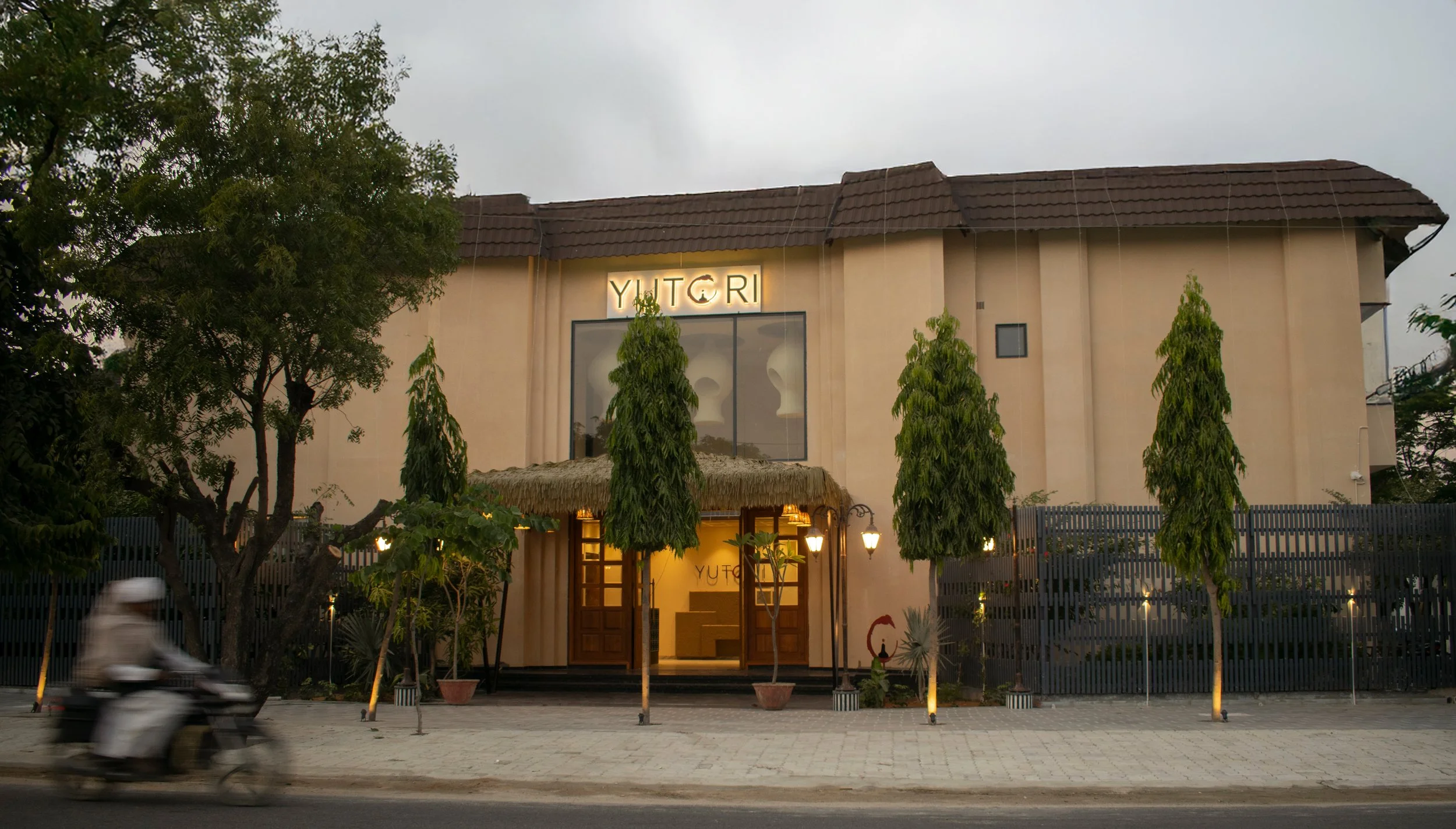 Exterior of a building with a sign that reads "YUTORI," tall trees in front, and a motorcycle on the street in the foreground.