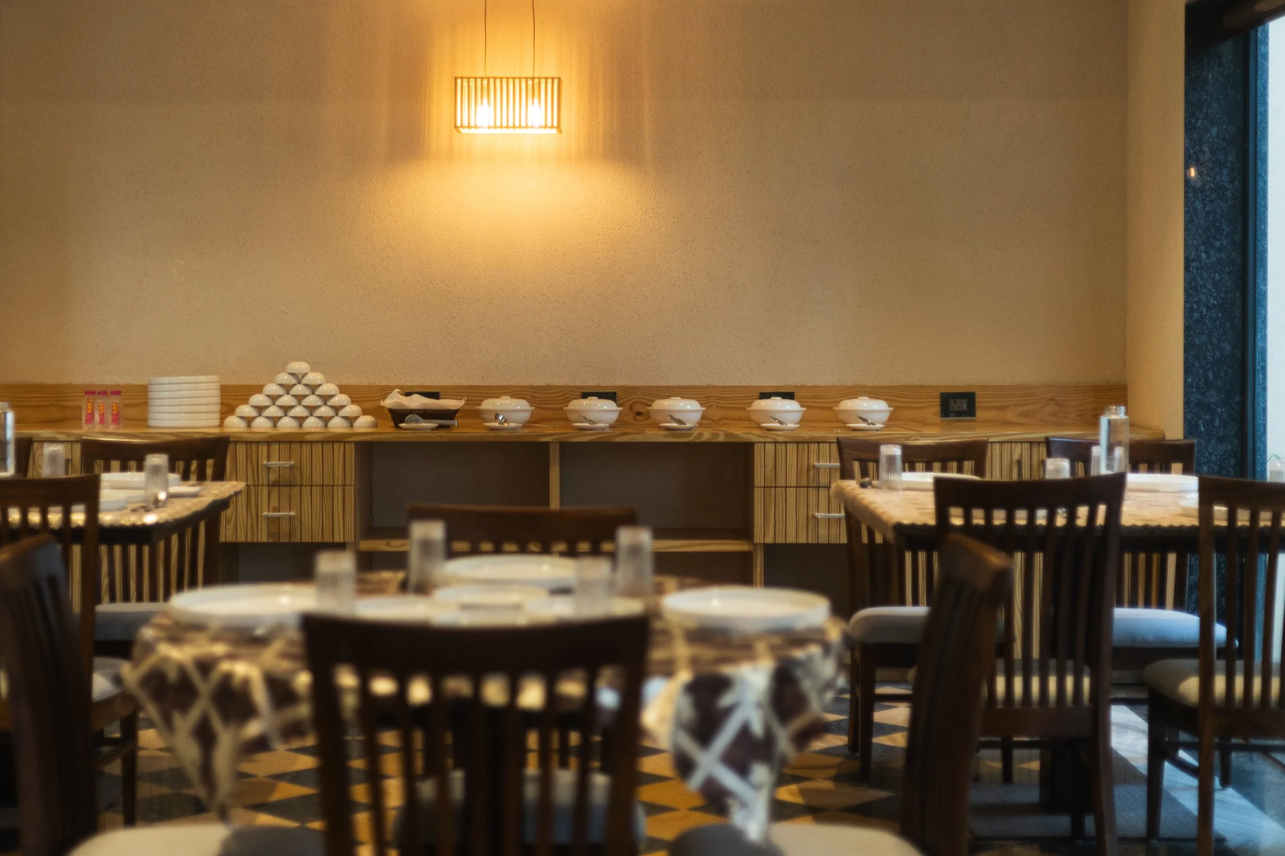 Empty restaurant with round tables covered with patterned tablecloths, set with glasses and plates, wooden chairs, and a buffet area with bowls and plates against a beige wall with a warm light fixture.