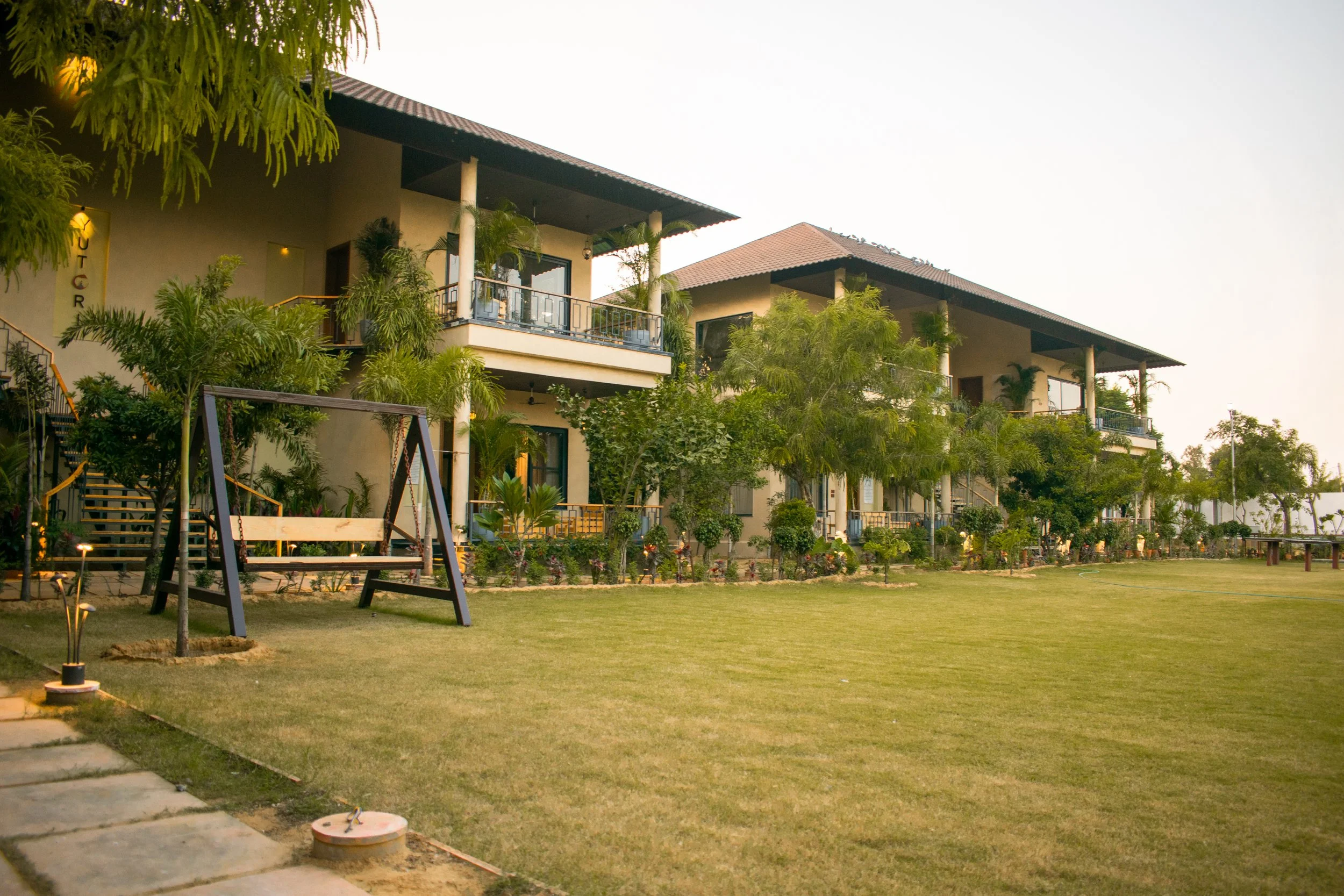 A large luxury villa or hotel with multiple floors, balconies, and a garden area with grass, trees, and outdoor furniture, during the daytime.