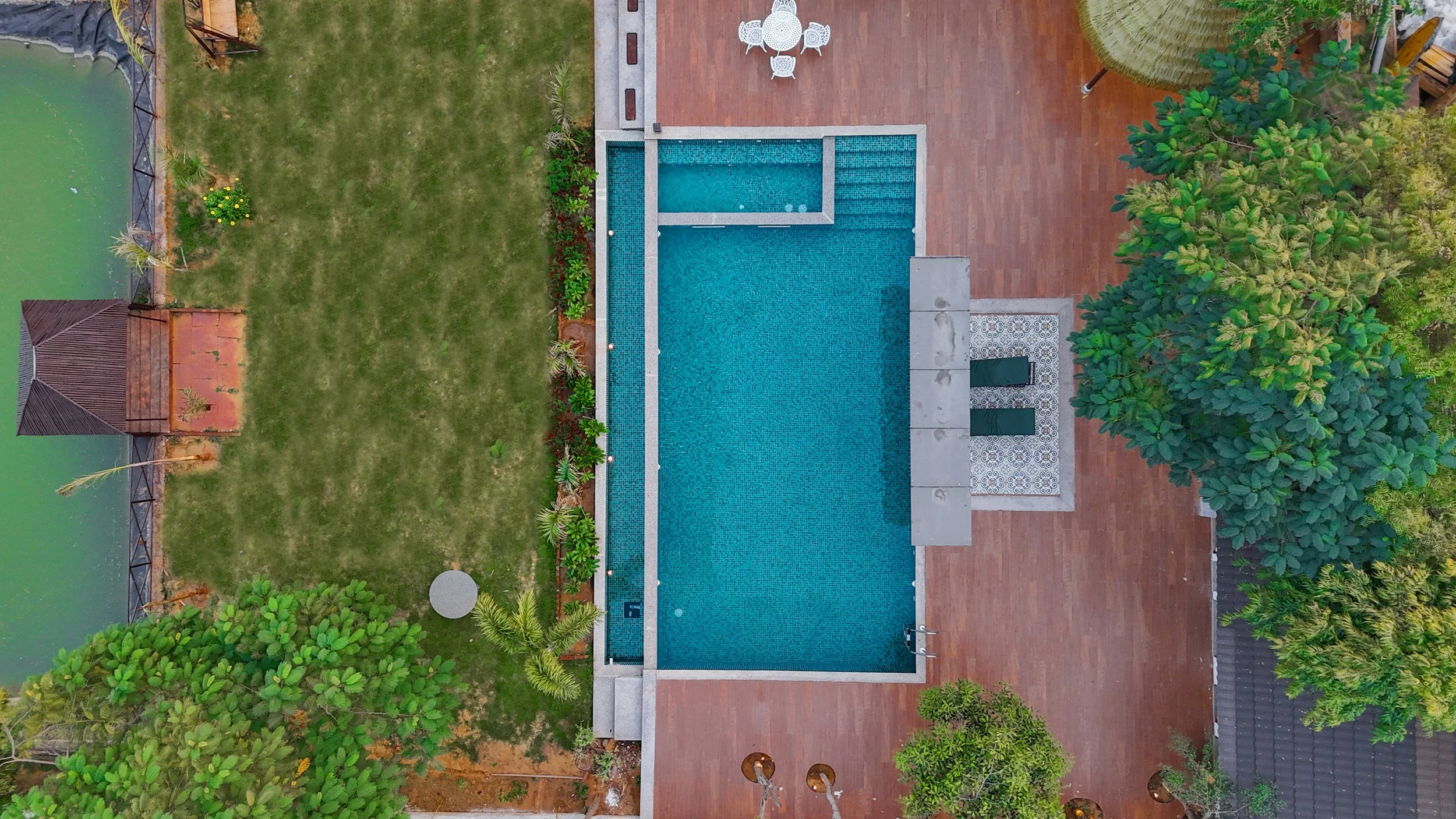 An aerial view of a backyard with a swimming pool, a brick patio, a grassy area, trees, and outdoor furniture.