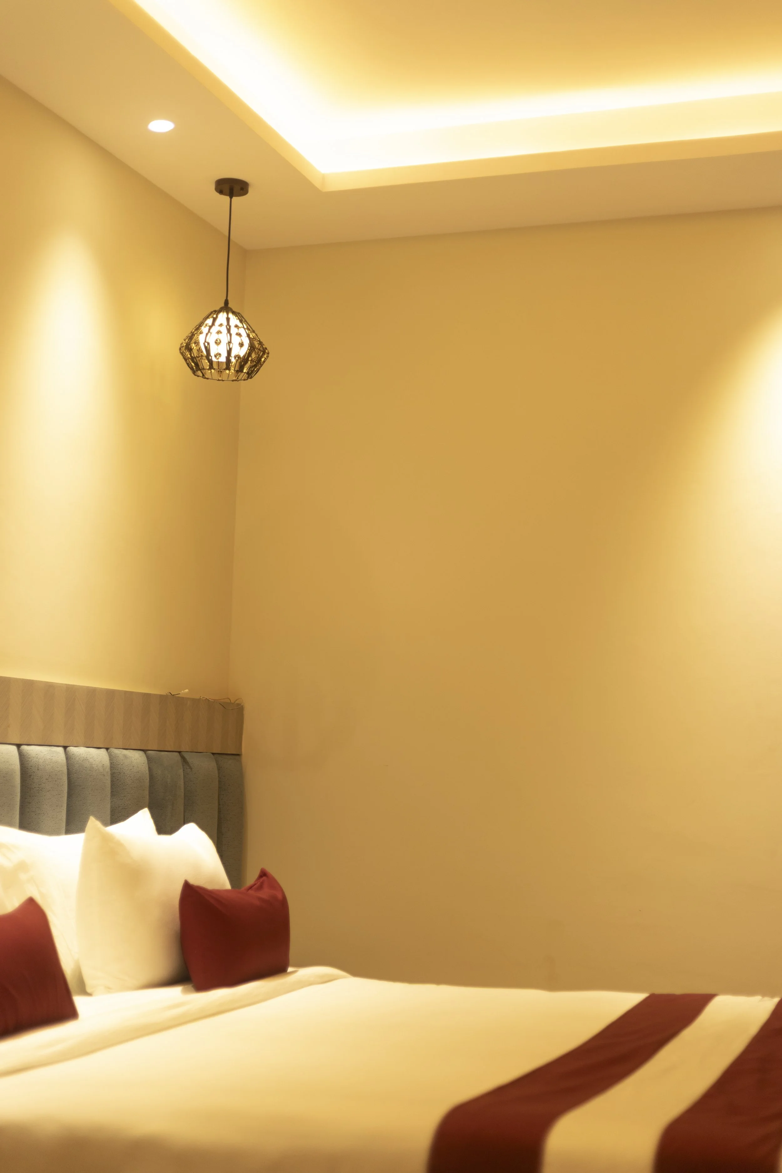 Close-up of a neatly made bed with white and red pillows in a warmly lit hotel room with yellow walls and a ceiling with hidden lighting