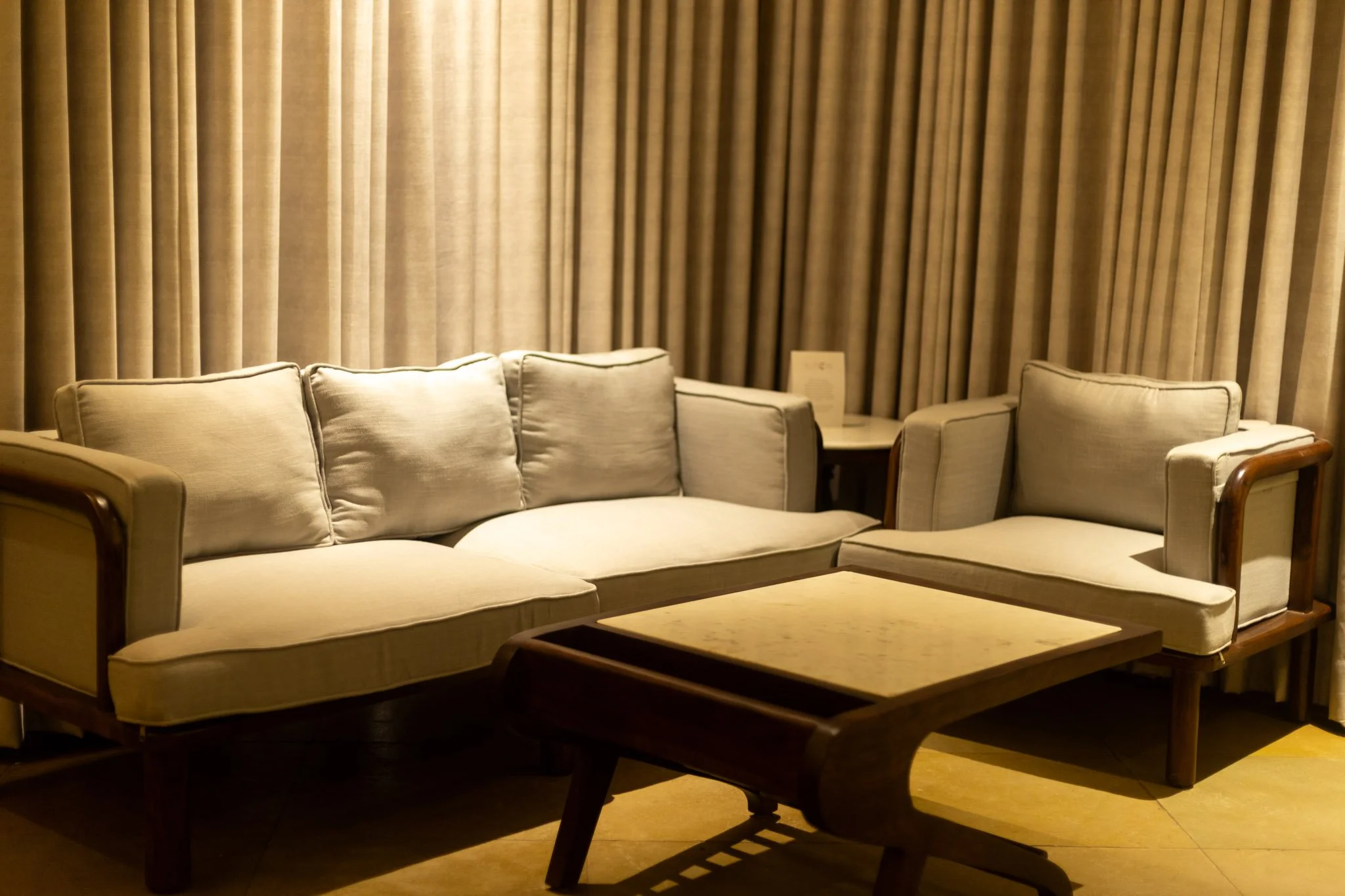 Living room with beige sofa, matching armchair, and wooden coffee table in front of a window with long beige curtains.