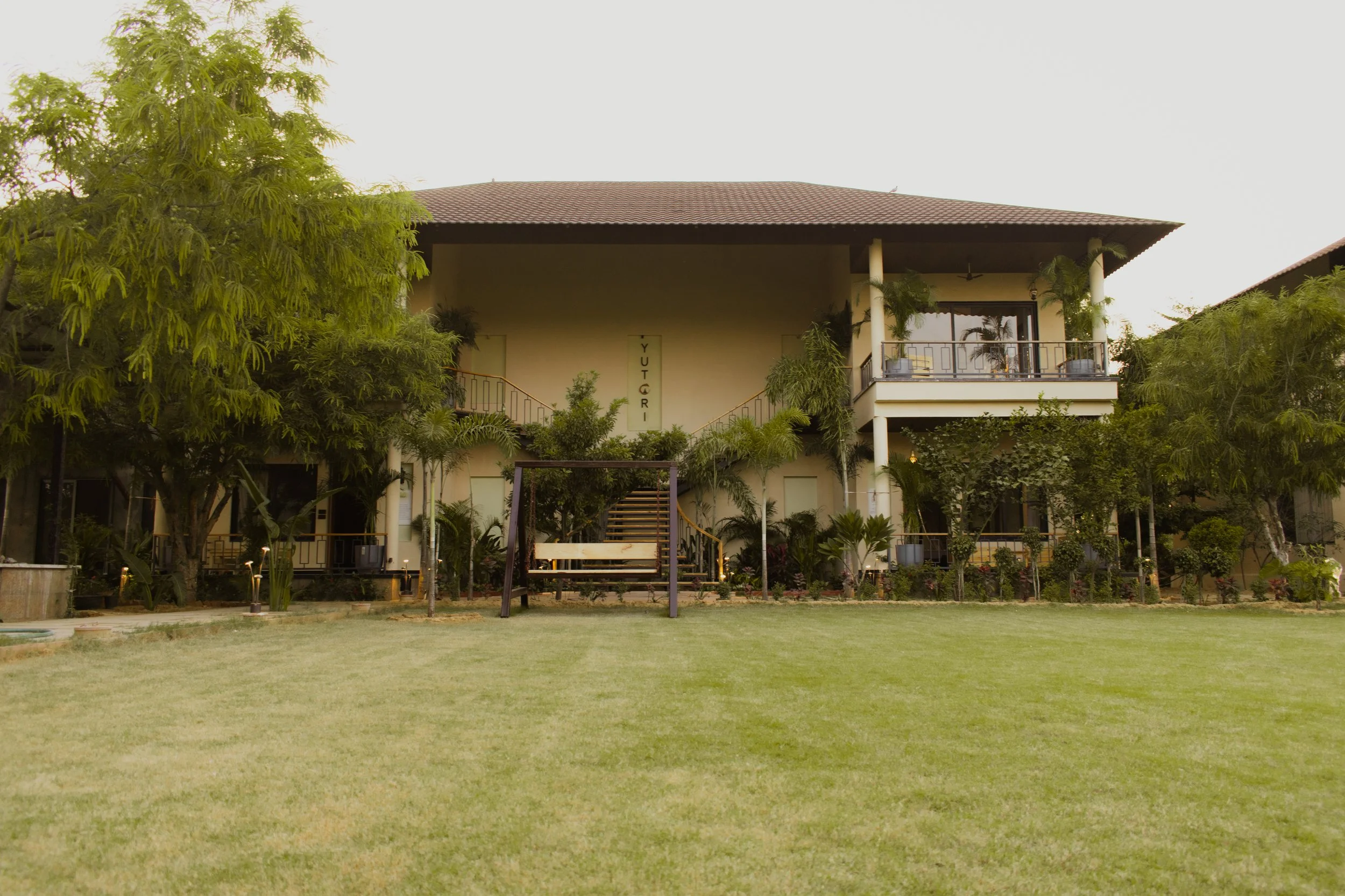 Large two-story building with a red-tiled roof and balconies, surrounded by lush green trees and a well-maintained lawn.