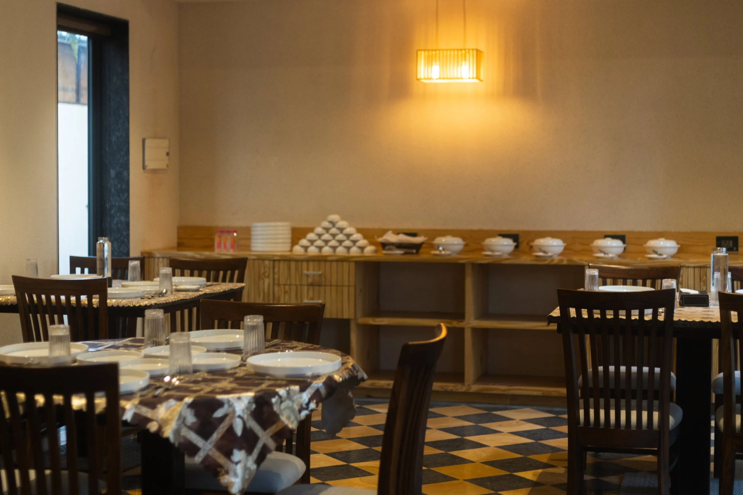 Restaurant dining area with tables set with plates, glasses, and utensils, and a buffet area with bowls and a pyramid of eggs on a wooden counter.