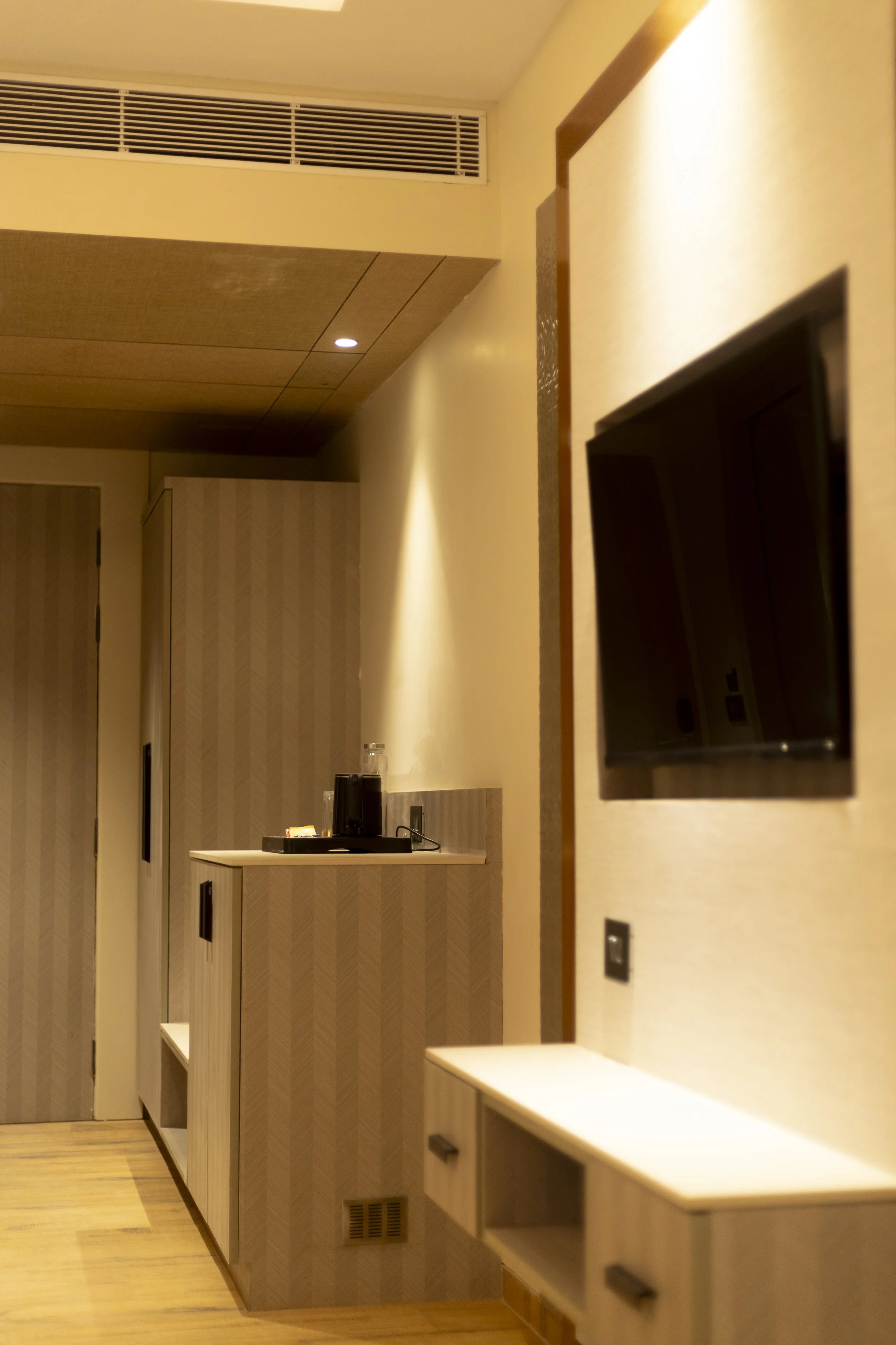 Interior of a hotel room with a small nook holding a coffee maker and supplies, a wall-mounted television, and a wooden cabinet, with wood flooring and beige walls.