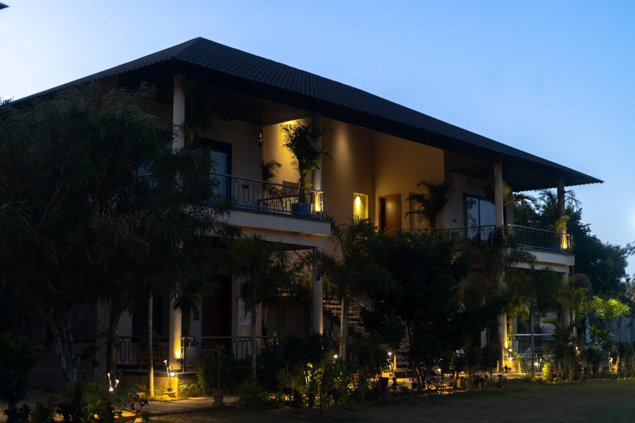 Two-story house with balconies, surrounded by trees, illuminated with exterior lights at dusk.