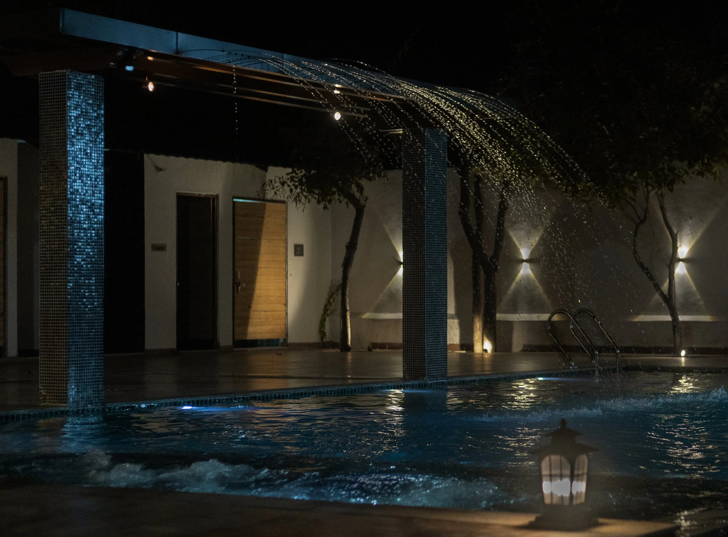 Night view of an outdoor swimming pool with a water feature that pours water from a canopy, surrounded by trees with lights, and a wooden door on a wall in the background.