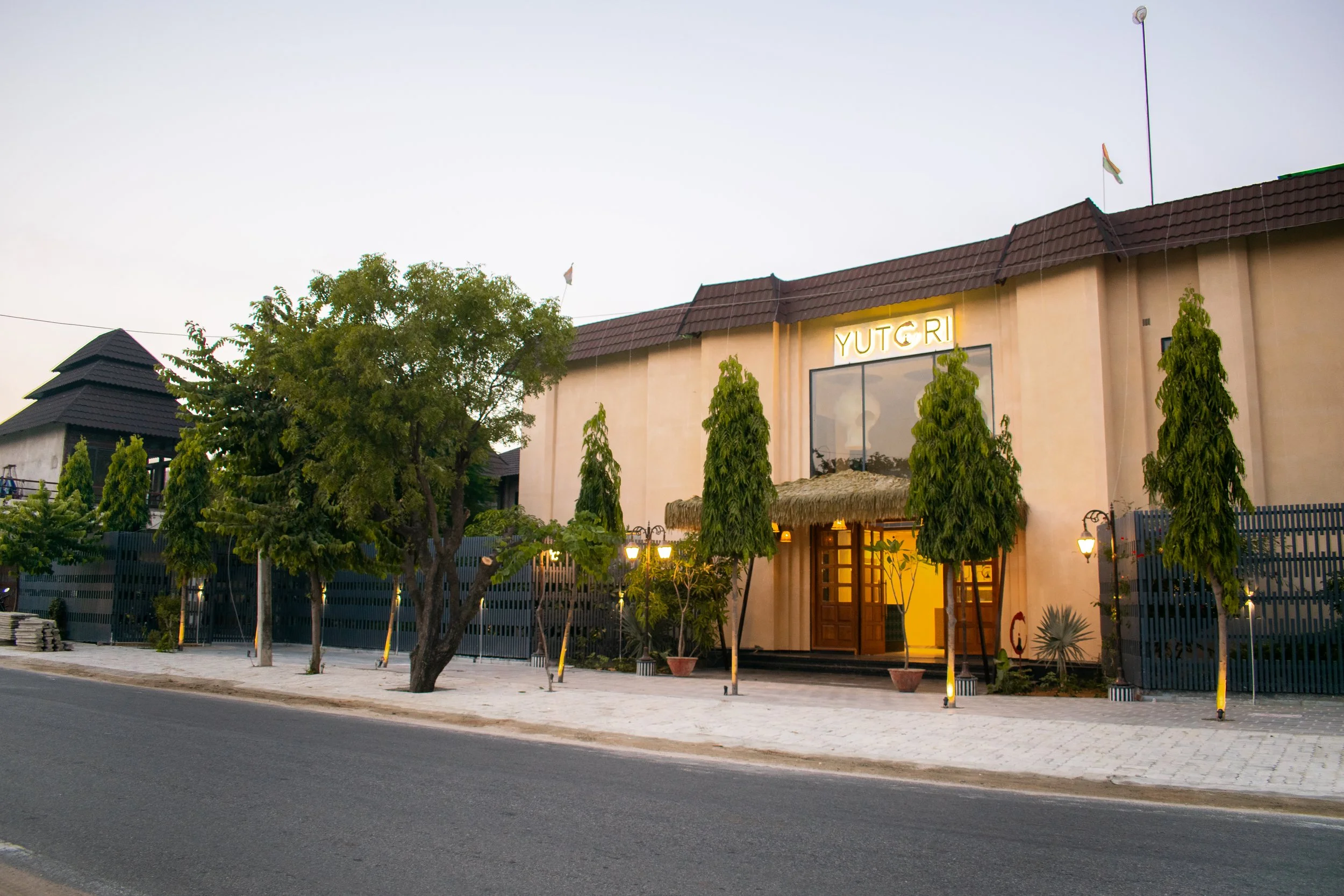 A two-story building with a sign saying 'YUTGRI' above a large window, surrounded by trees and street lights on a paved sidewalk, taken during early evening.