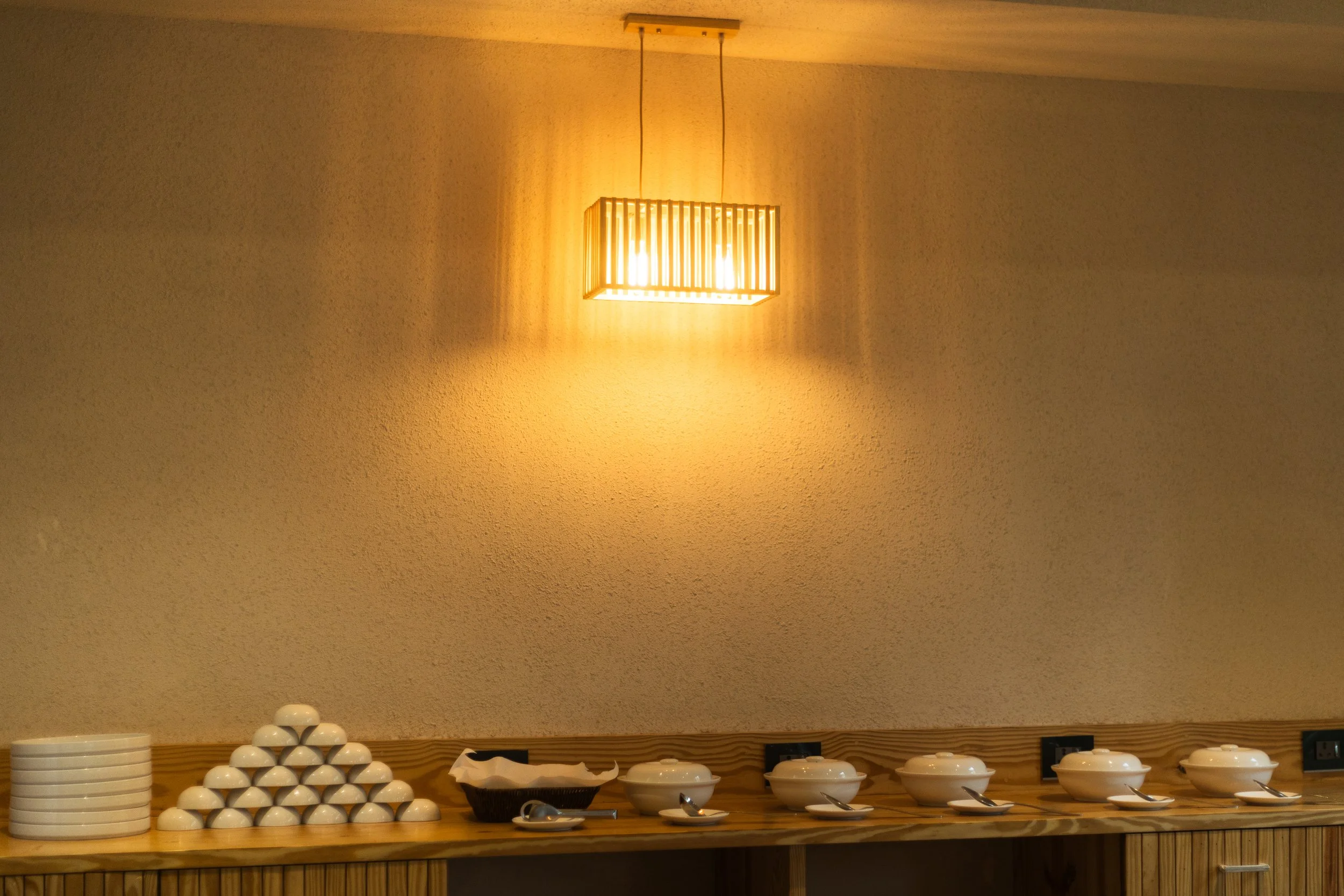 A buffet table with white bowls, plates and serving dishes, on a wooden counter against a textured beige wall illuminated by a yellow pendant lamp.