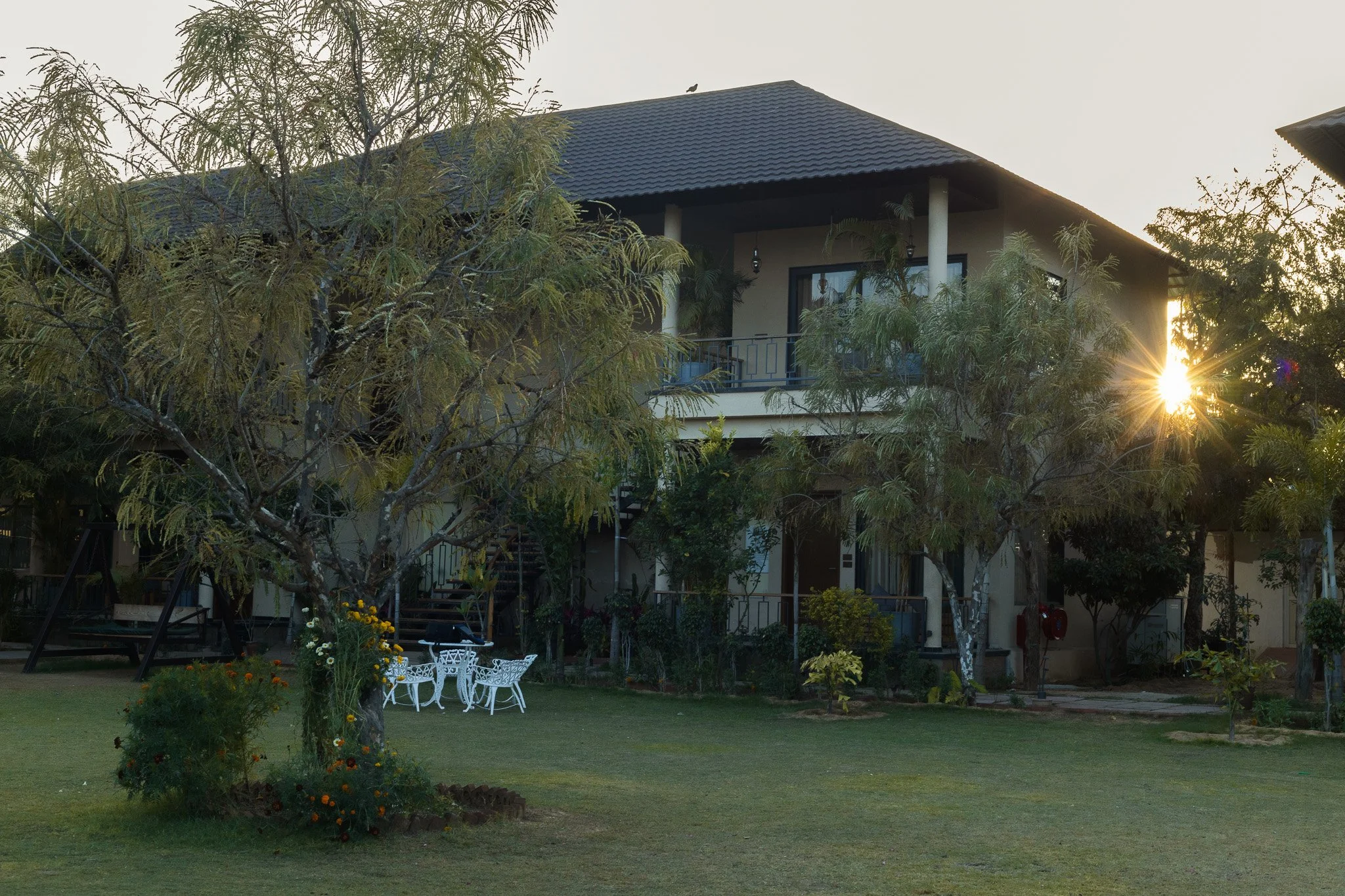 A two-story house with a dark roof, surrounded by trees and a garden, with the setting sun shining near the roof.