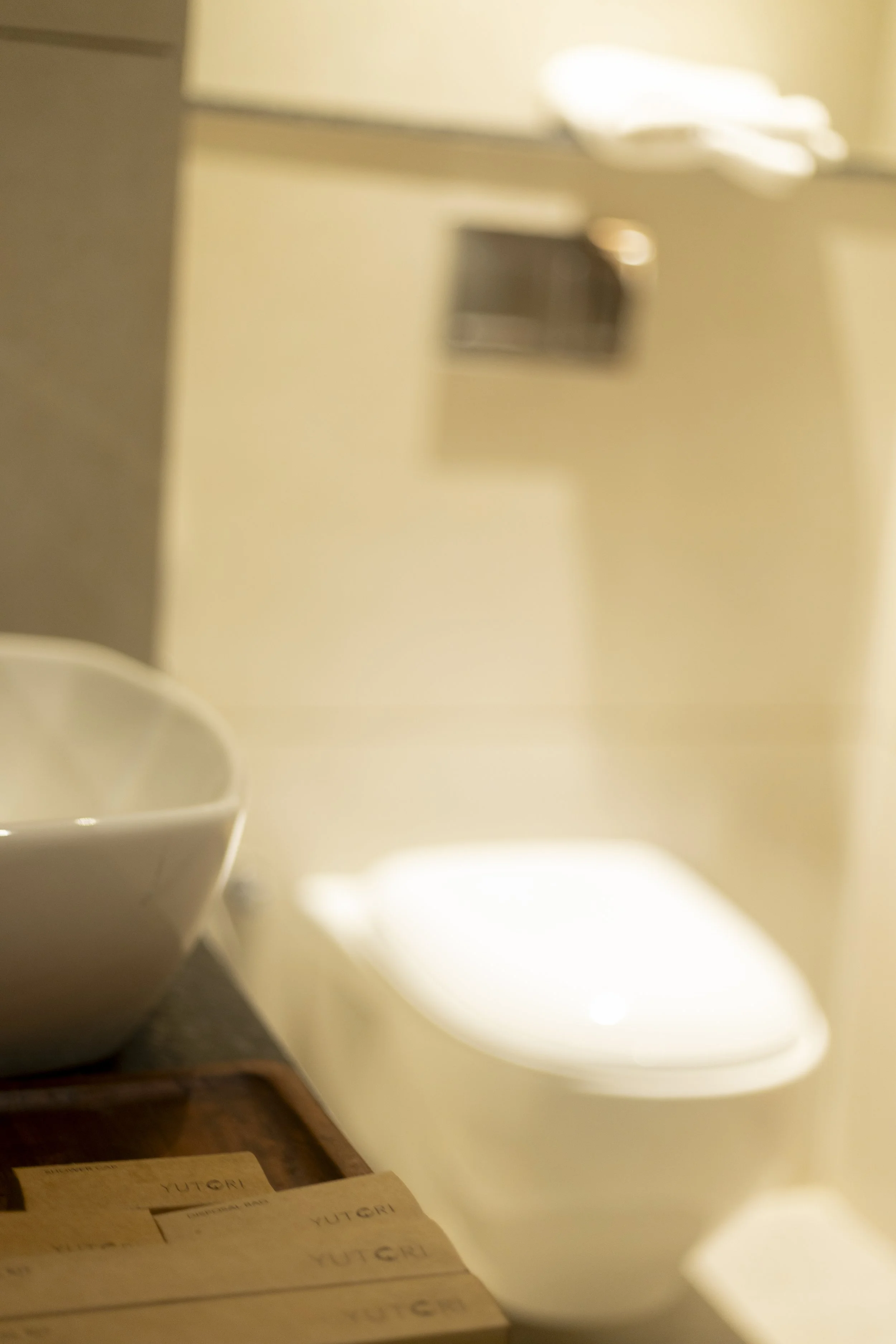 A bathroom with a white toilet, a white bowl, and a wooden surface with papers labeled 'YUTGRI' in the foreground. The background shows a beige wall with a framed picture and a towel on top.
