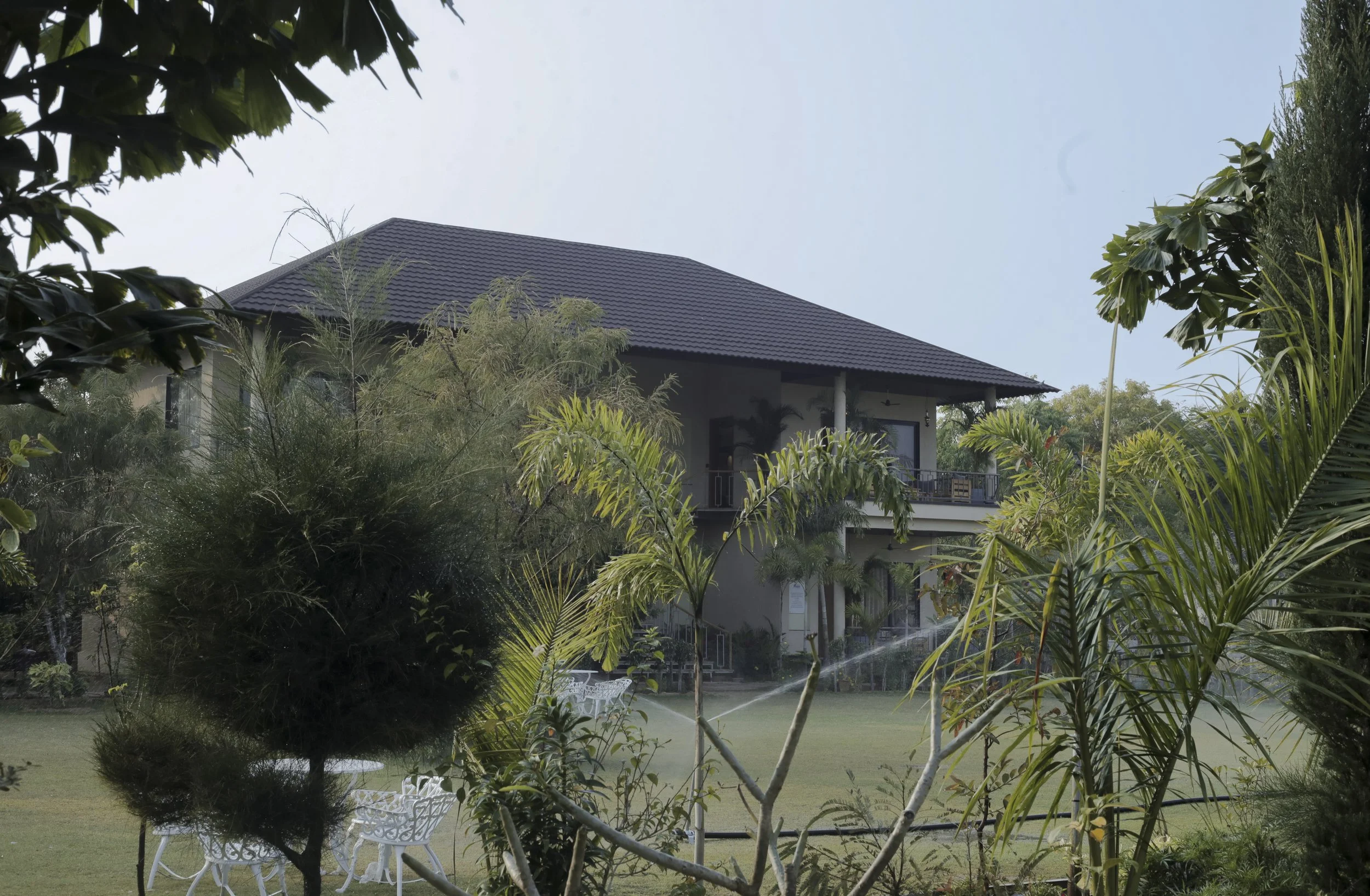 A two-story house with a tiled roof, surrounded by lush greenery and palm trees, with a sprinkler watering the lawn in the foreground.
