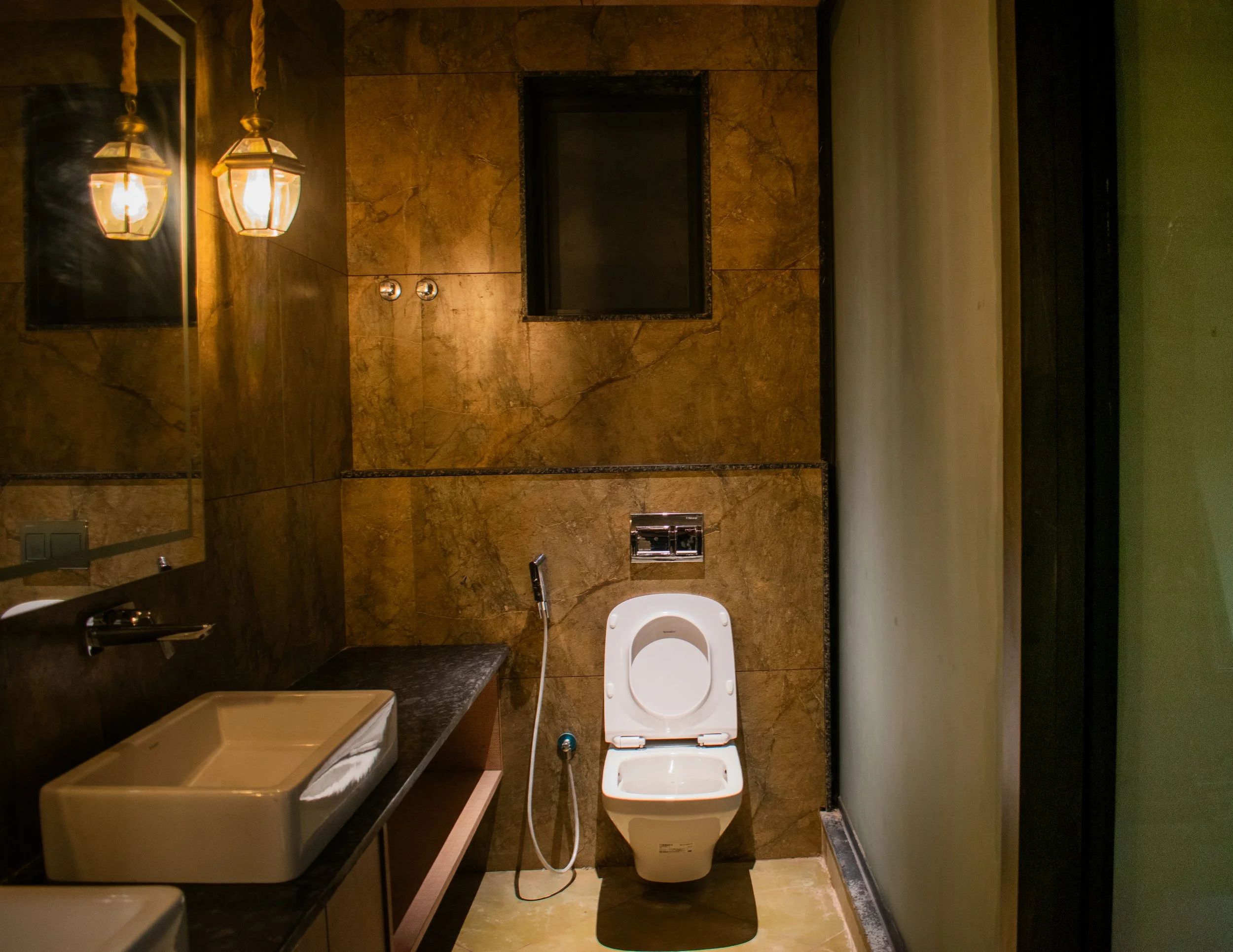 Modern bathroom with brown marble tiles, a white toilet, a beige stone sink, a black countertop, and two pendant lights hanging above the mirror.