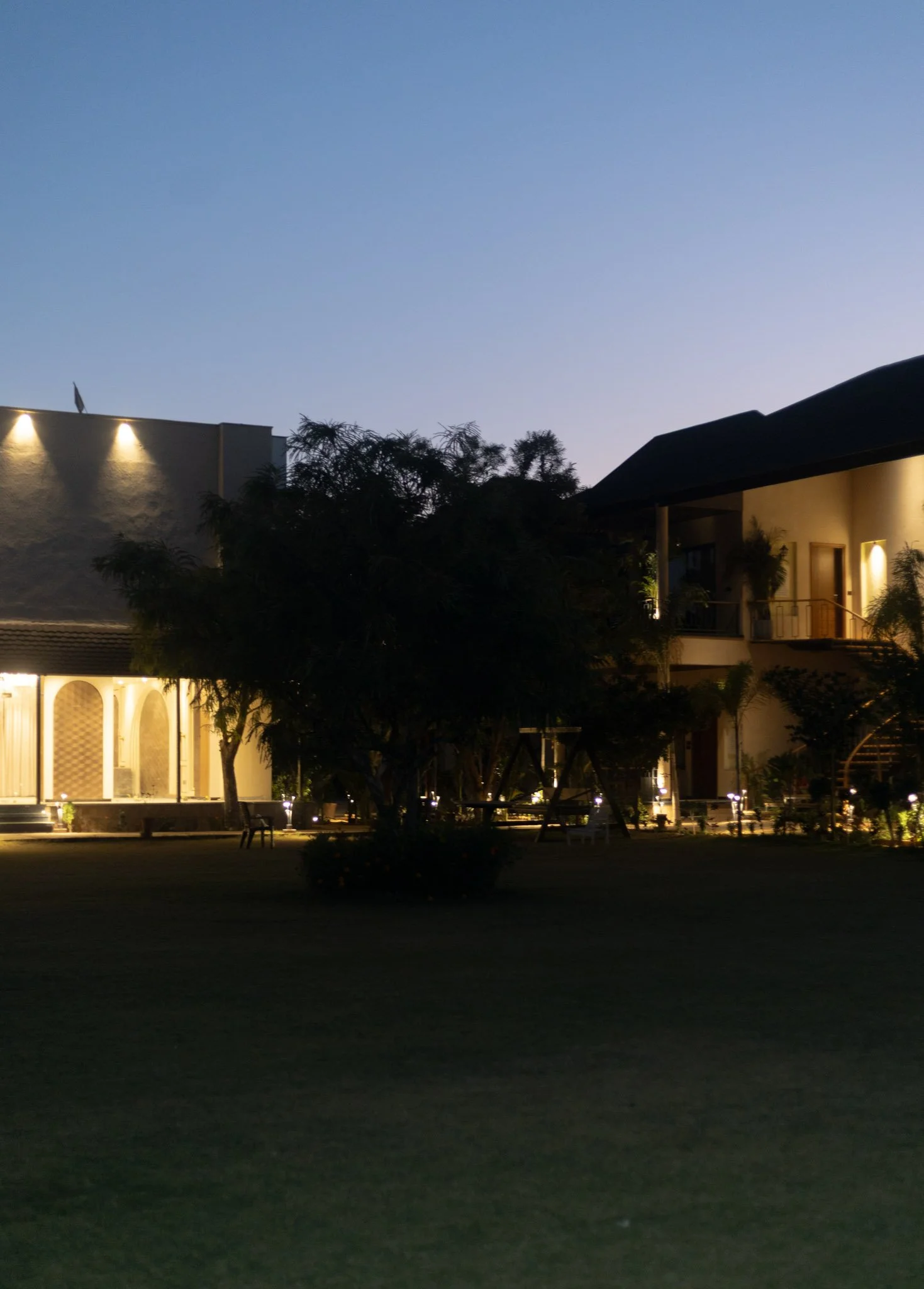 Nighttime scene of a modern house with exterior lights, trees, and a playground with a swing set outside.