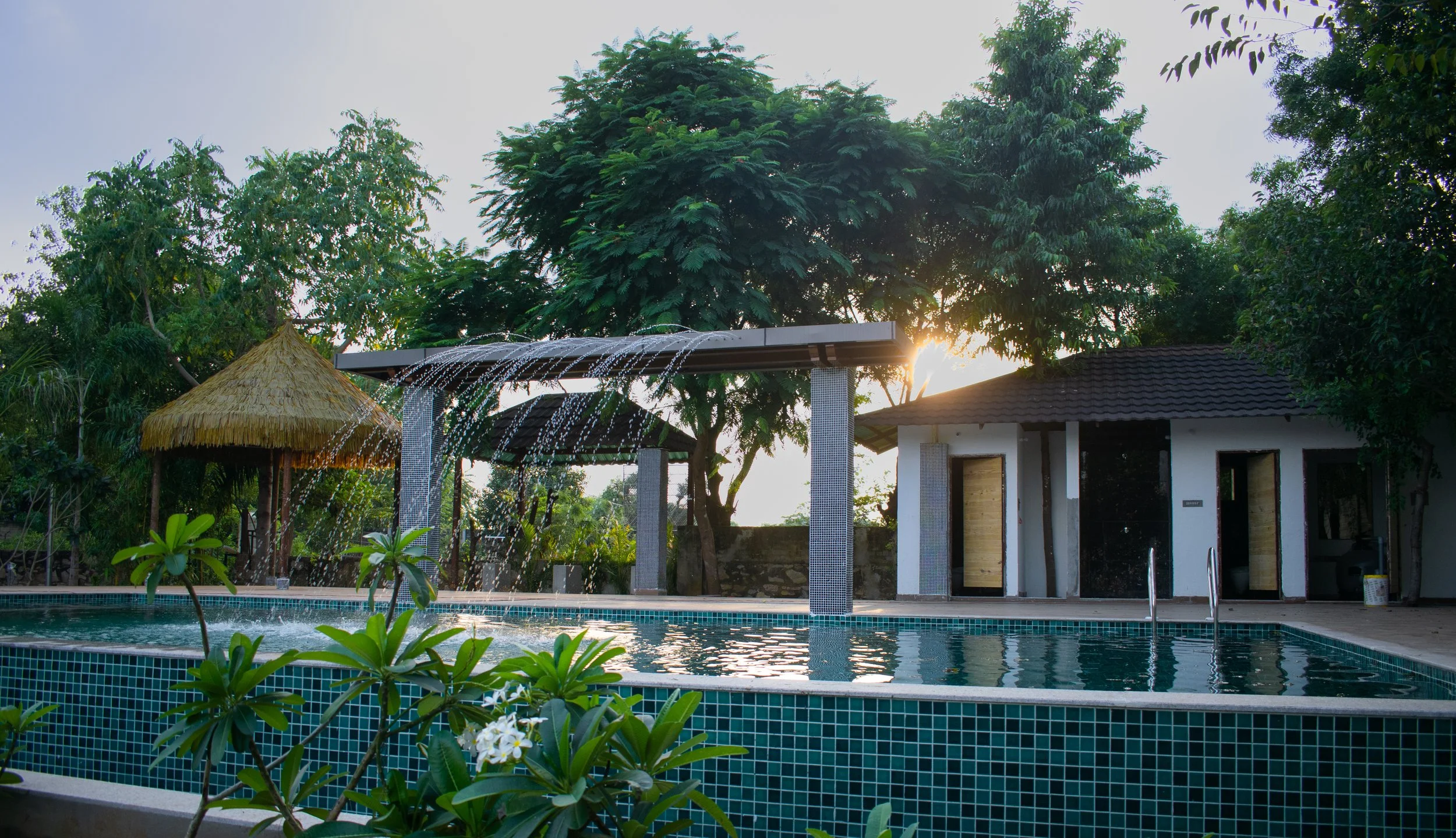 Swimming pool with small waterfall feature, surrounded by lush green trees and plants, with a thatched hut and a small building in the background during sunset.