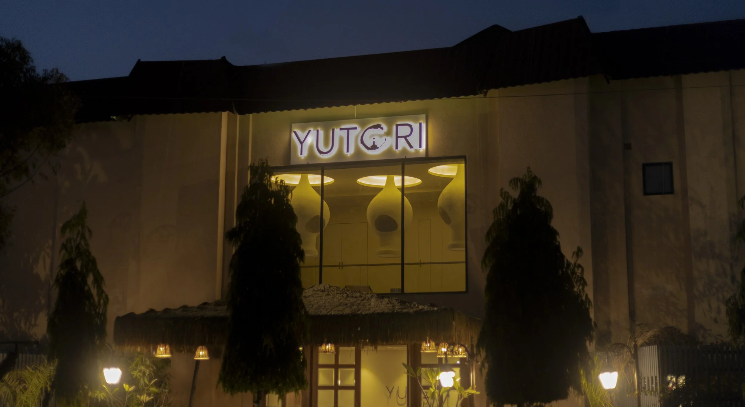 Night view of a building with a sign that reads "YUTORI" illuminated above a large window, showing hanging lamps inside. Trees and outdoor lighting are visible in the foreground.