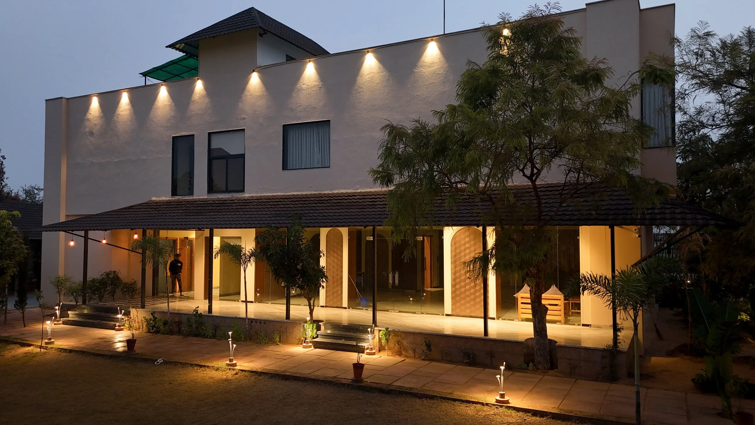 A modern multi-story house at dusk with exterior lights, a porch, and surrounding trees and plants.