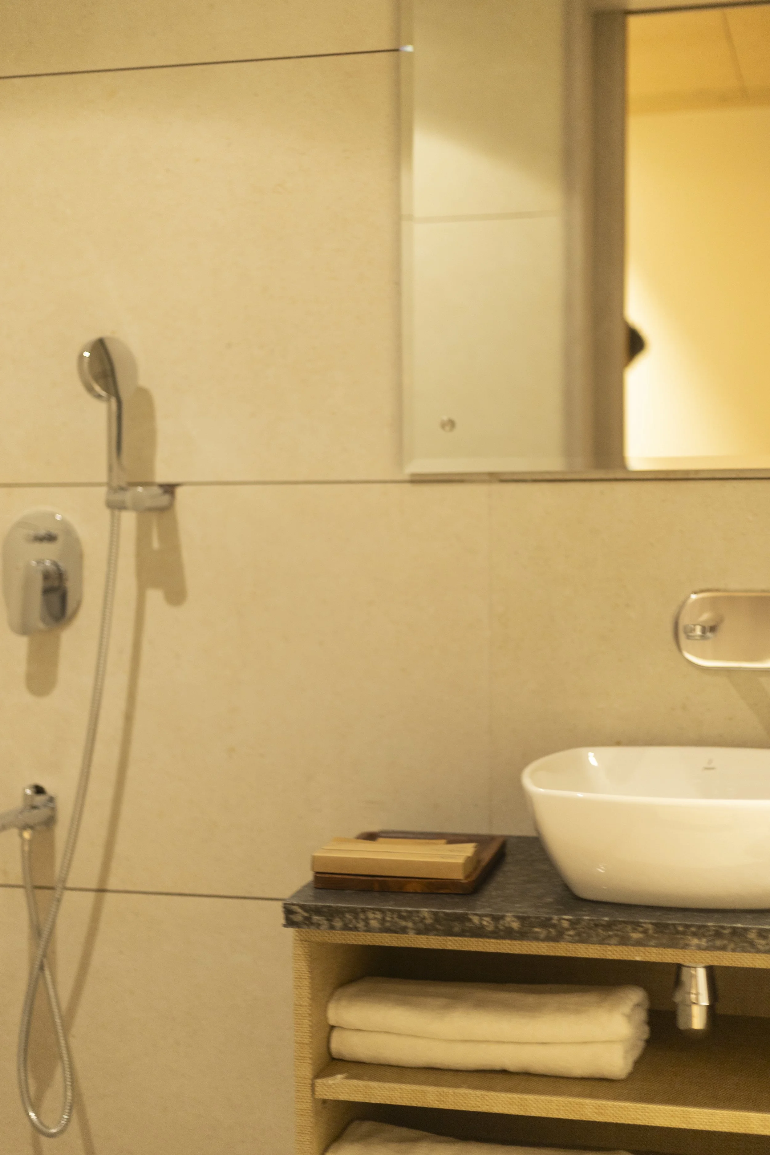 A bathroom with a showerhead on the left, a mirror above a sink on the right, a countertop with a windowed cabinet underneath, and towels stored on a lower shelf.