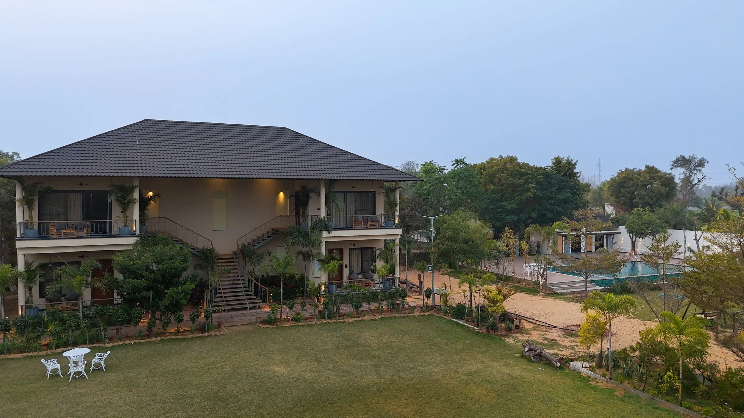 Two-story house with balconies, surrounded by greenery and trees, with a garden, outdoor seating, and a swimming pool in the background.