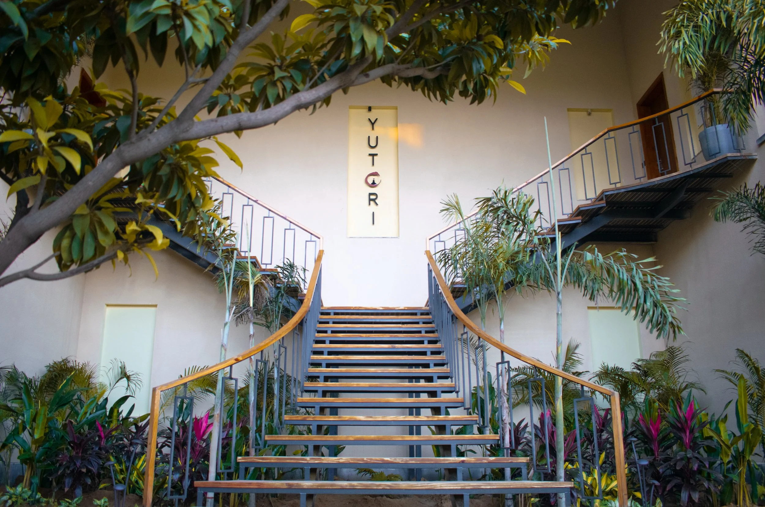 Exterior view of a staircase leading to a second floor with plants and trees around. A vertical sign with the text 'YUTCRI' is visible on the wall, which is cream-colored. The staircase has black railings and wooden handrails, with the surrounding ar