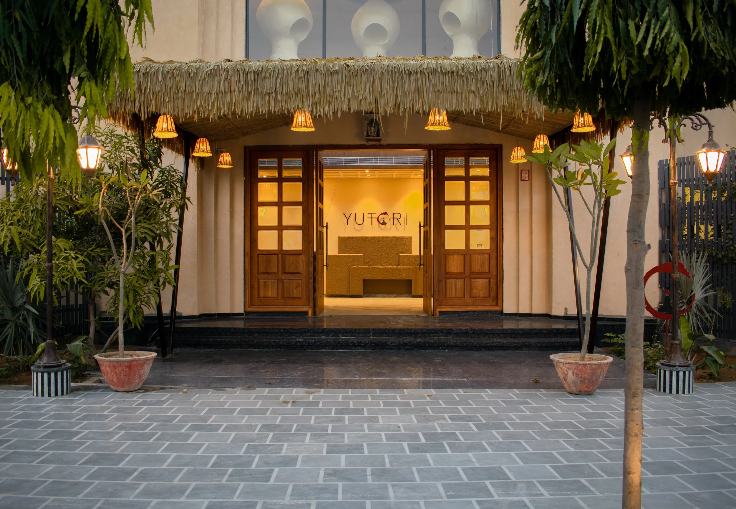 Entrance of a building with wooden doors and warm lighting, surrounded by plants, with a sign that reads "YUTORI" in the background.