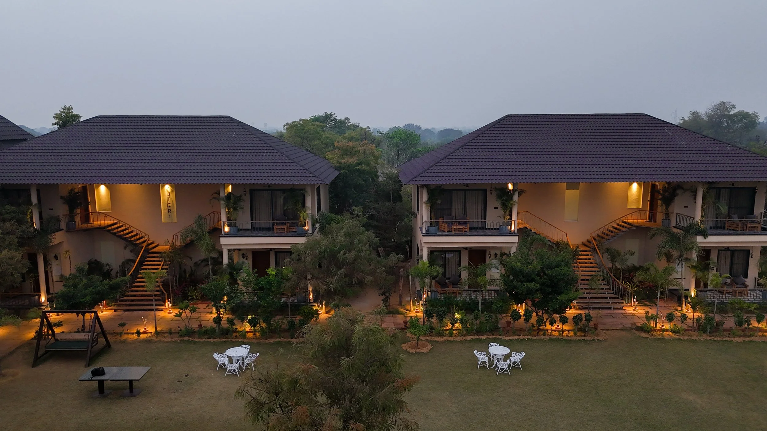 Two modern two-story buildings with dark tiled roofs, exterior lighting, balconies, and staircases in a lush landscaped area at dusk, with trees, grass, and outdoor seating.