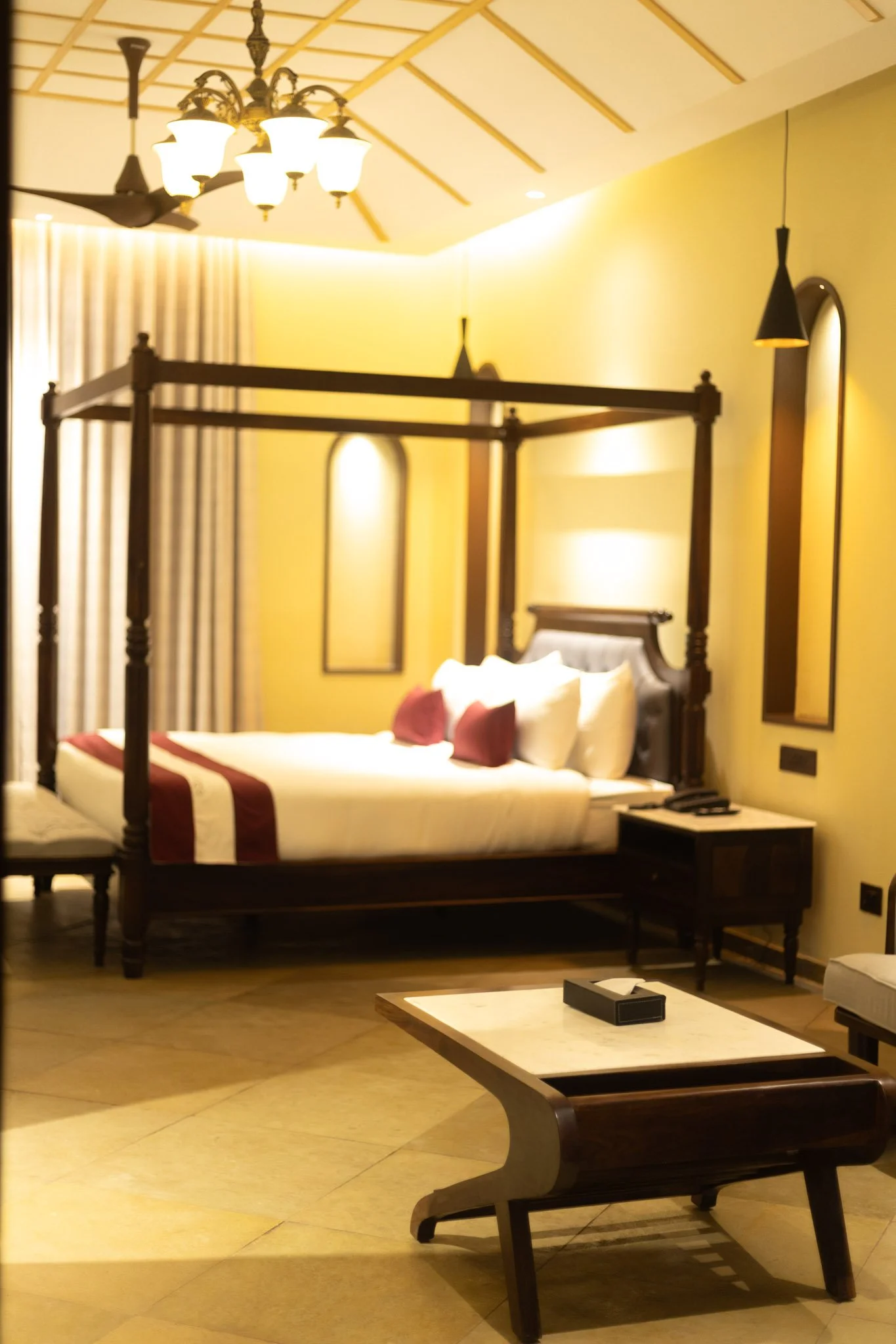 A hotel room with a four-poster bed, decorative lighting, and a central table.