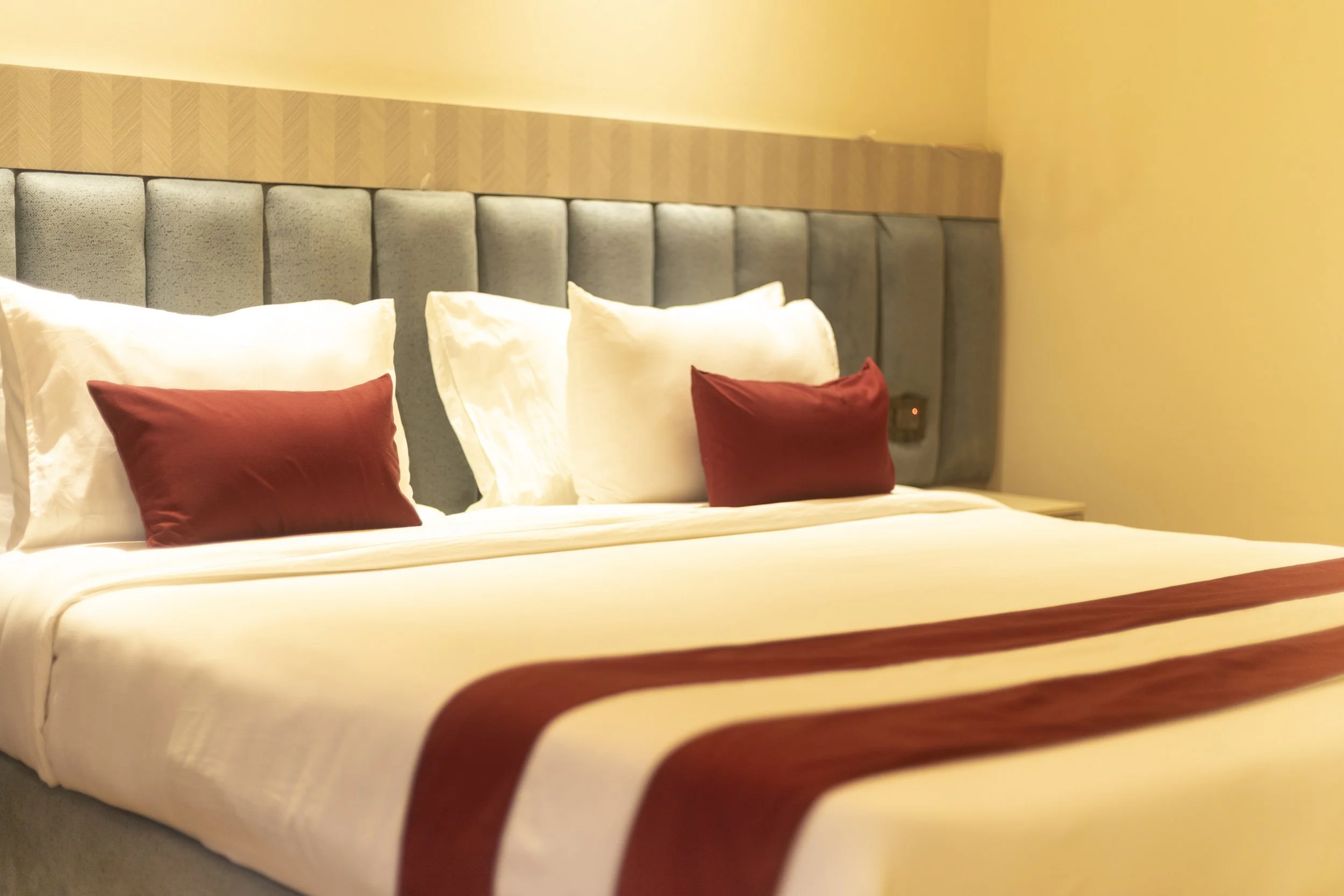 A neatly made hotel bed with white linens, two white pillows, two red decorative pillows, and a red bed runner, with a grey upholstered headboard and a beige wall in the background.