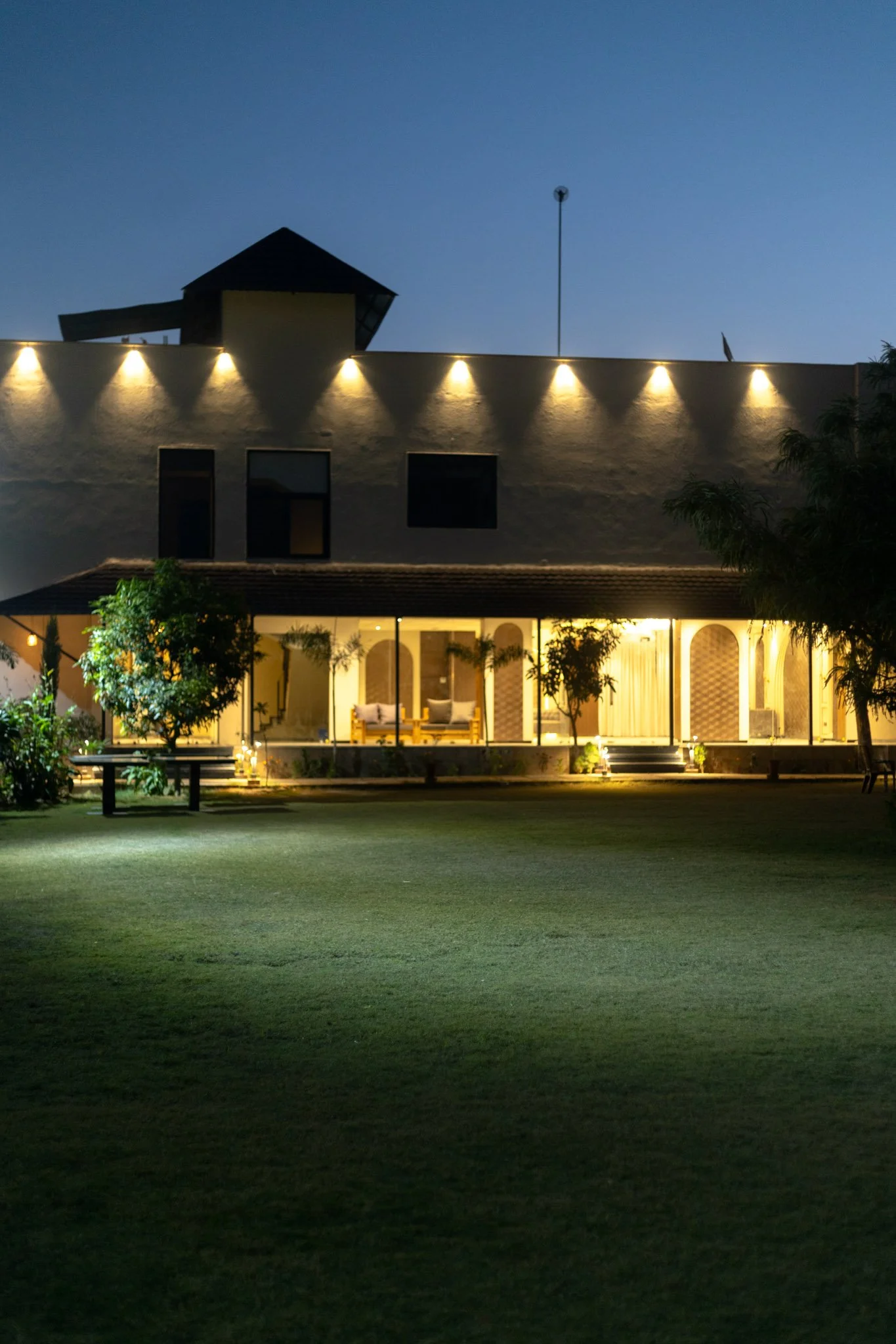 A nighttime view of a modern house with large glass windows, illuminated from within, surrounded by a green lawn and trees, with exterior lights on the upper part of the building and a clear evening sky in the background.
