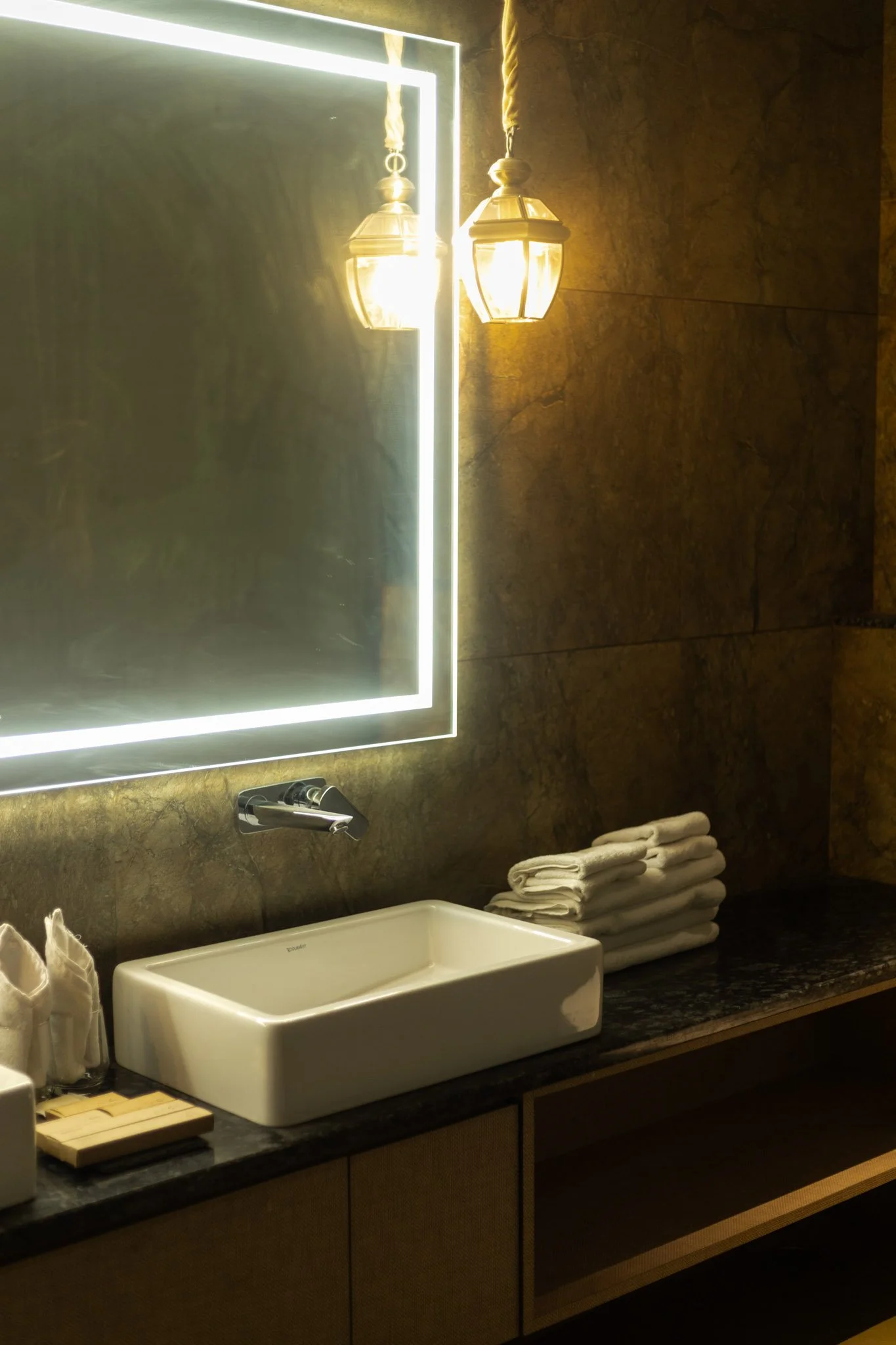 A bathroom sink area with a rectangular white vessel sink, a mirrored wall with a bright border, two decorative hanging lamps, a stack of folded white towels, soap, and toiletries on a dark countertop with a textured wall background.