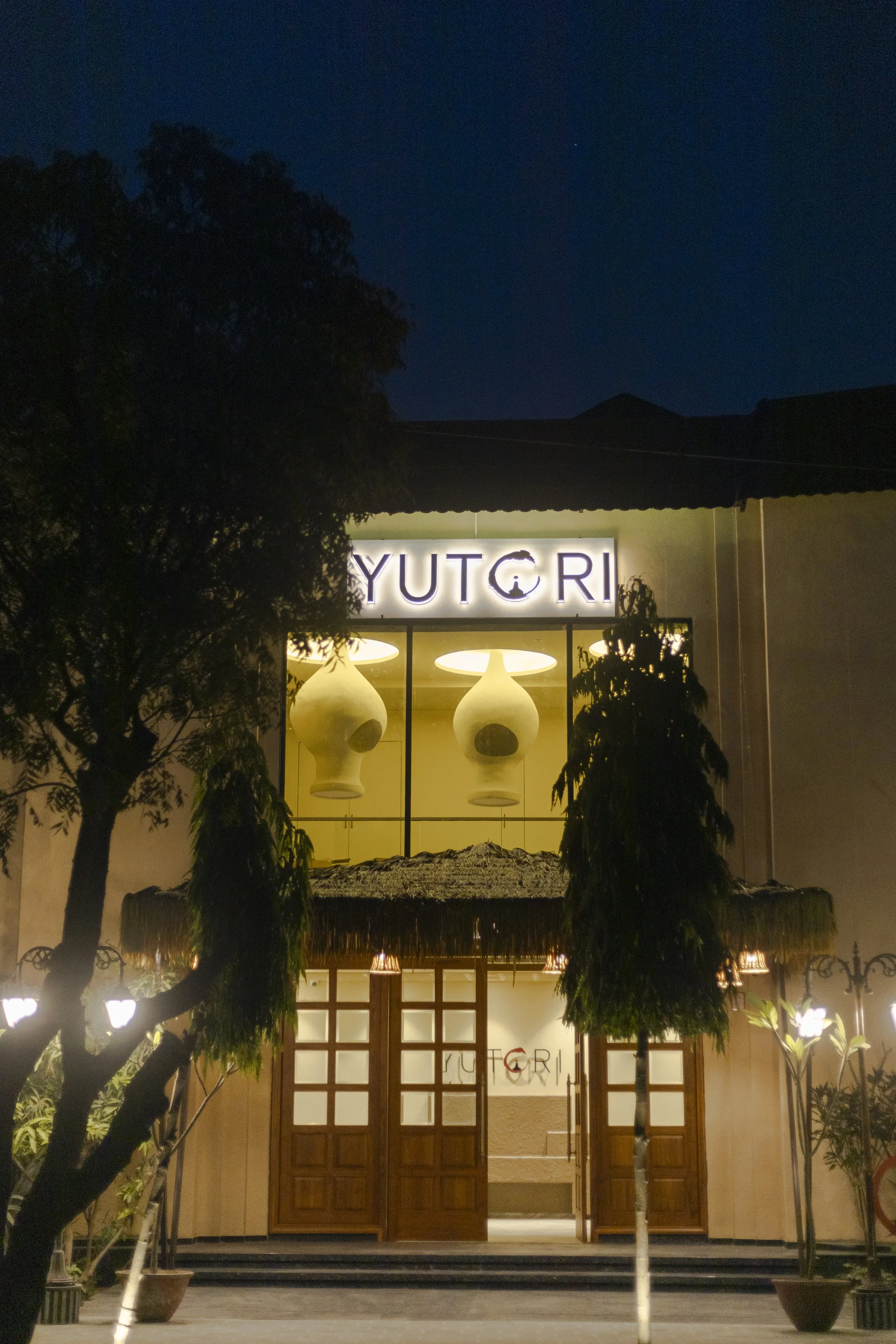 Night view of a restaurant named Yutgri with a wooden entrance and decorative hanging lamps inside, surrounded by trees and outdoor lighting.