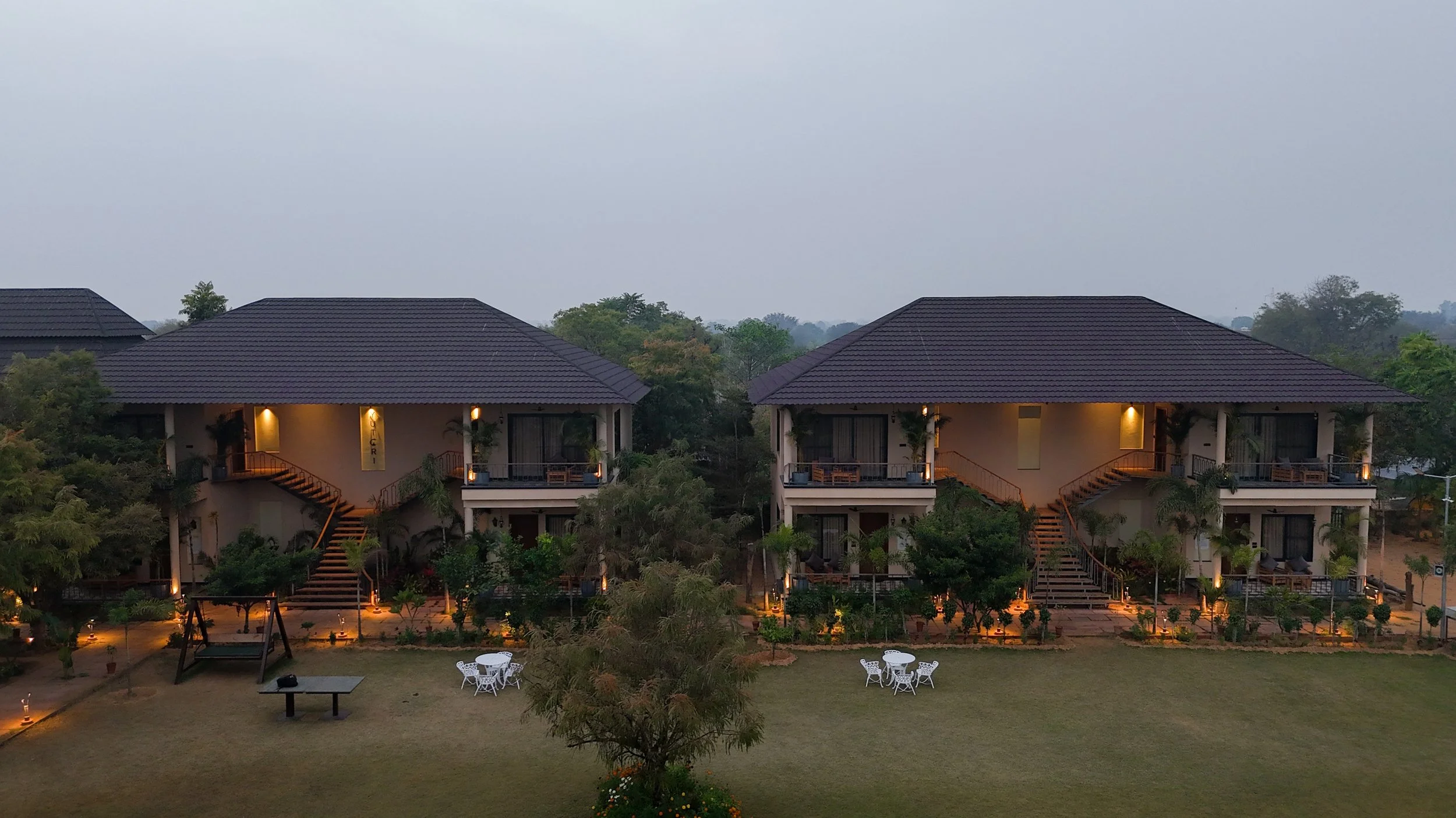 Nighttime scene of a modern house with exterior lights, trees, and a playground with a swing set outside.