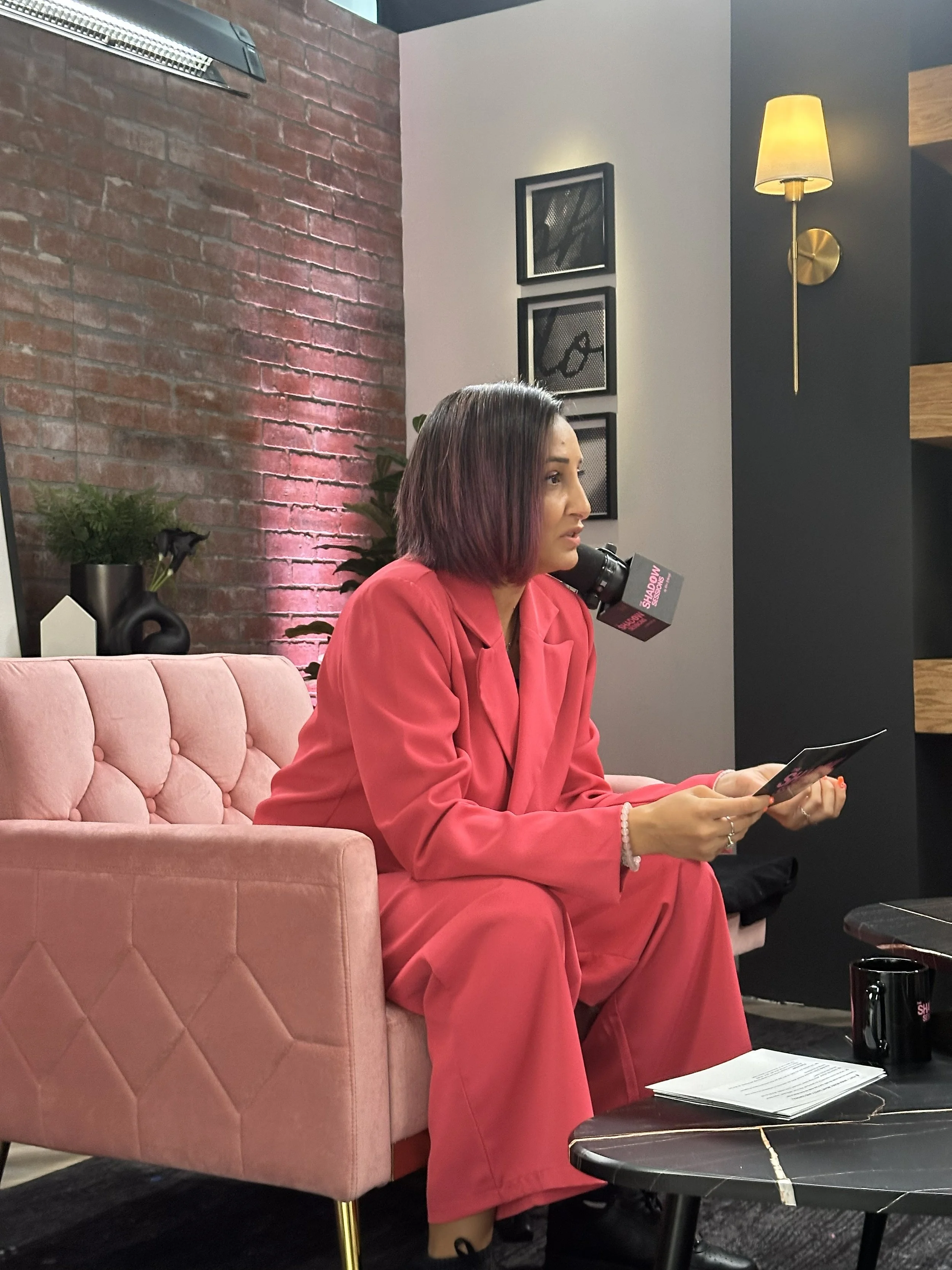 A woman in a pink suit sitting on a pink tufted armchair, reading a pamphlet, with a microphone labeled "Shadow" positioned near her mouth, indoors with a brick wall, framed pictures, plants, a lamp, and a black coffee mug on a black marble table.