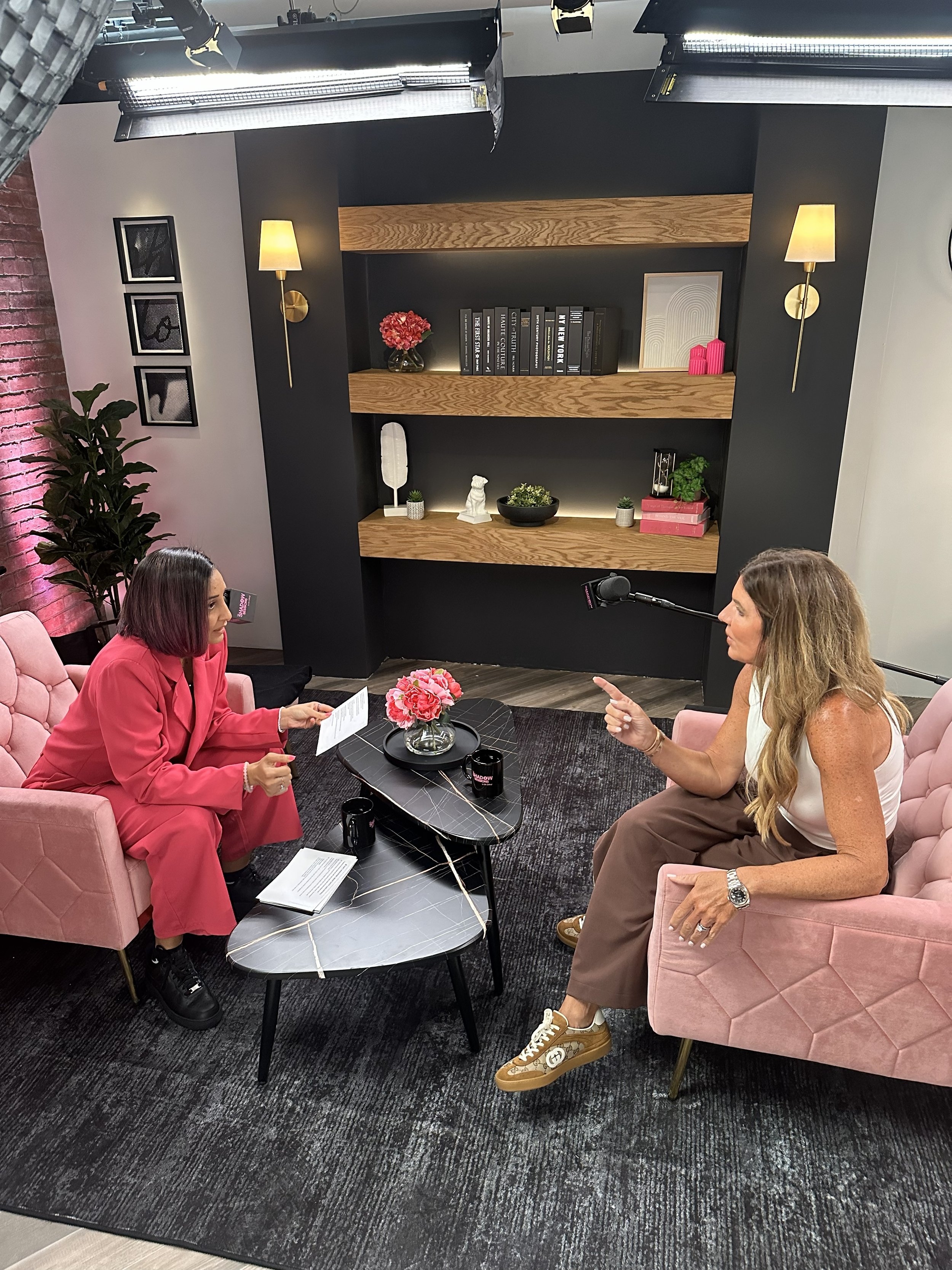 Two women engaged in a conversation in a cozy, modern studio with black and pink decor. One woman in a pink suit sits on a pink sofa, holding papers. The other woman in a white sleeveless top and brown pants sits on a matching pink sofa. There is a black marble coffee table with pink flowers and black mugs between them, and a black bookshelf with decorative items on the wall behind them.
