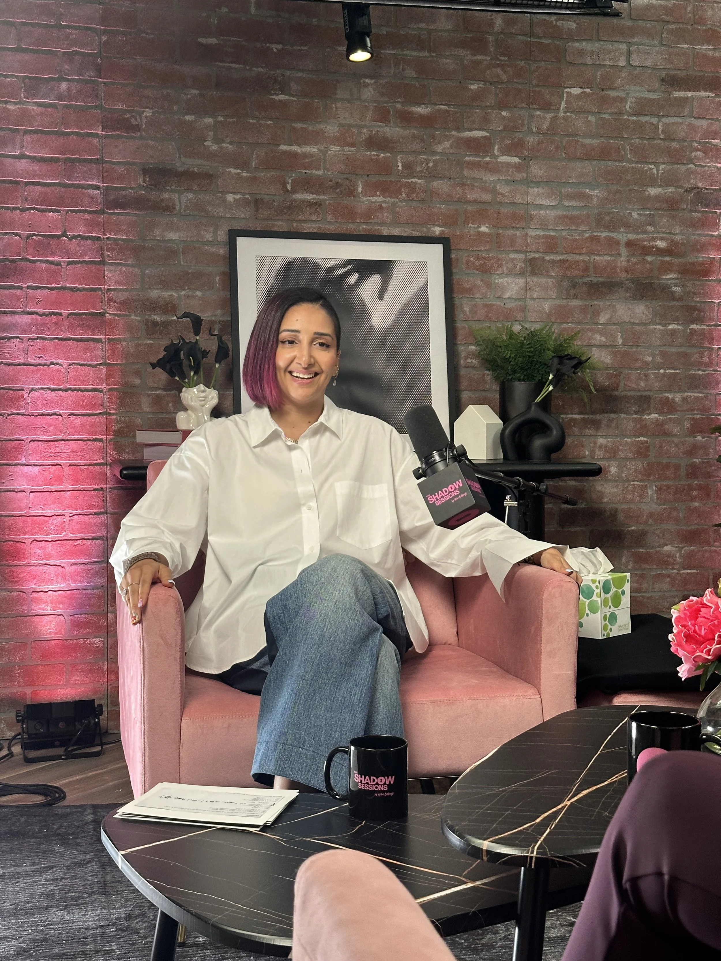 Woman with dark hair and pink tips sitting on a pink velvet armchair, smiling, in front of a microphone labeled 'Shadow Sessions,' with a brick wall, framed black and white art, and decorative plants in the background.