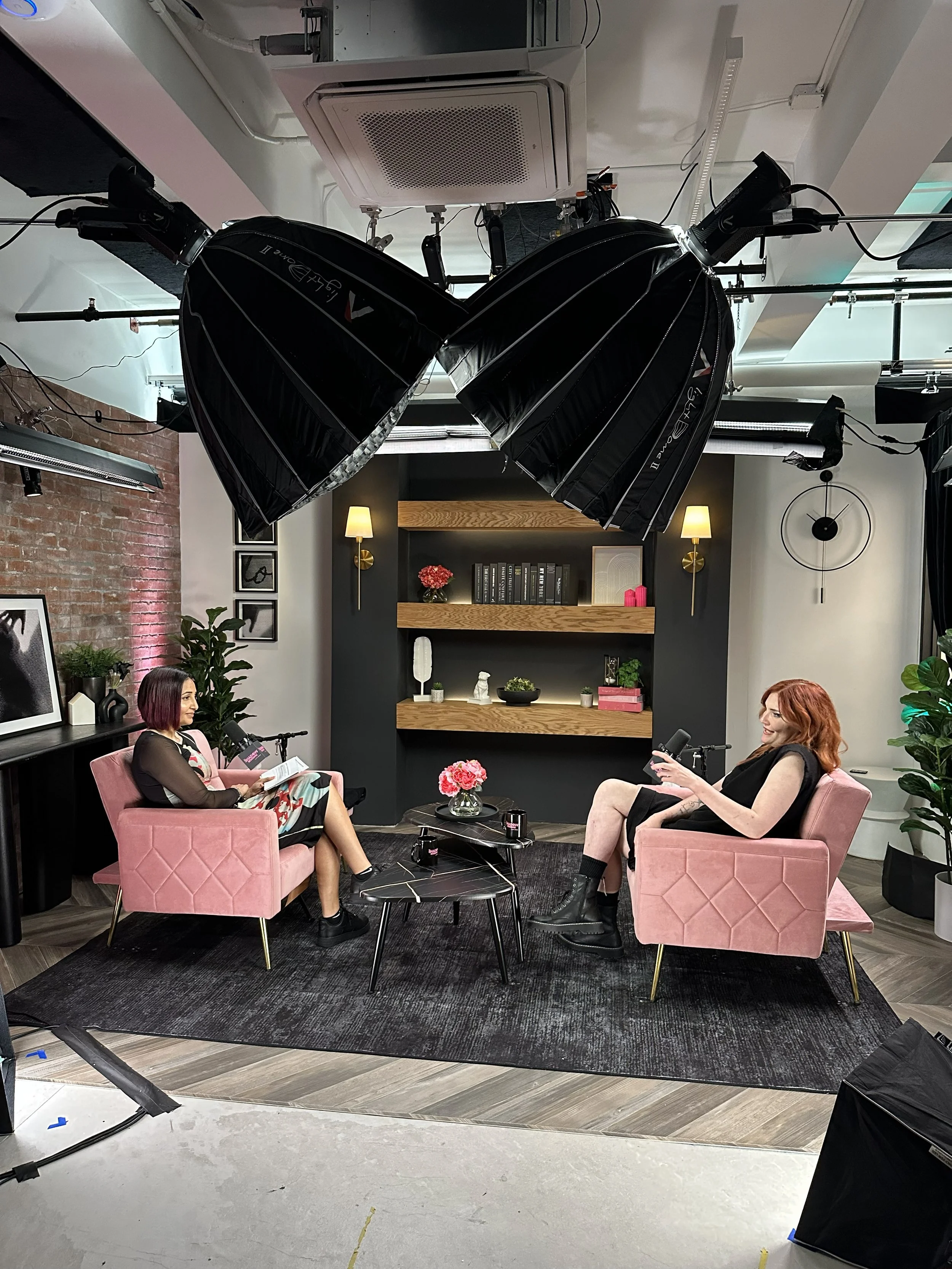 Two women are sitting in pink armchairs in a TV studio, engaged in conversation during an interview, with a black rug and a small black coffee table with flowers between them, surrounded by professional lighting equipment.