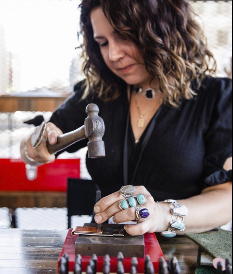 Ashley Vick working with jewelry, using a hammer over a small piece on a red cushion, wearing multiple rings and bracelets.