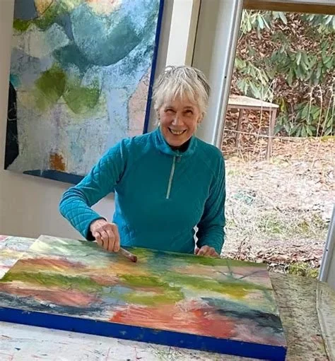 Carmen Grier smiling while painting a colorful landscape on a large canvas in an art studio.