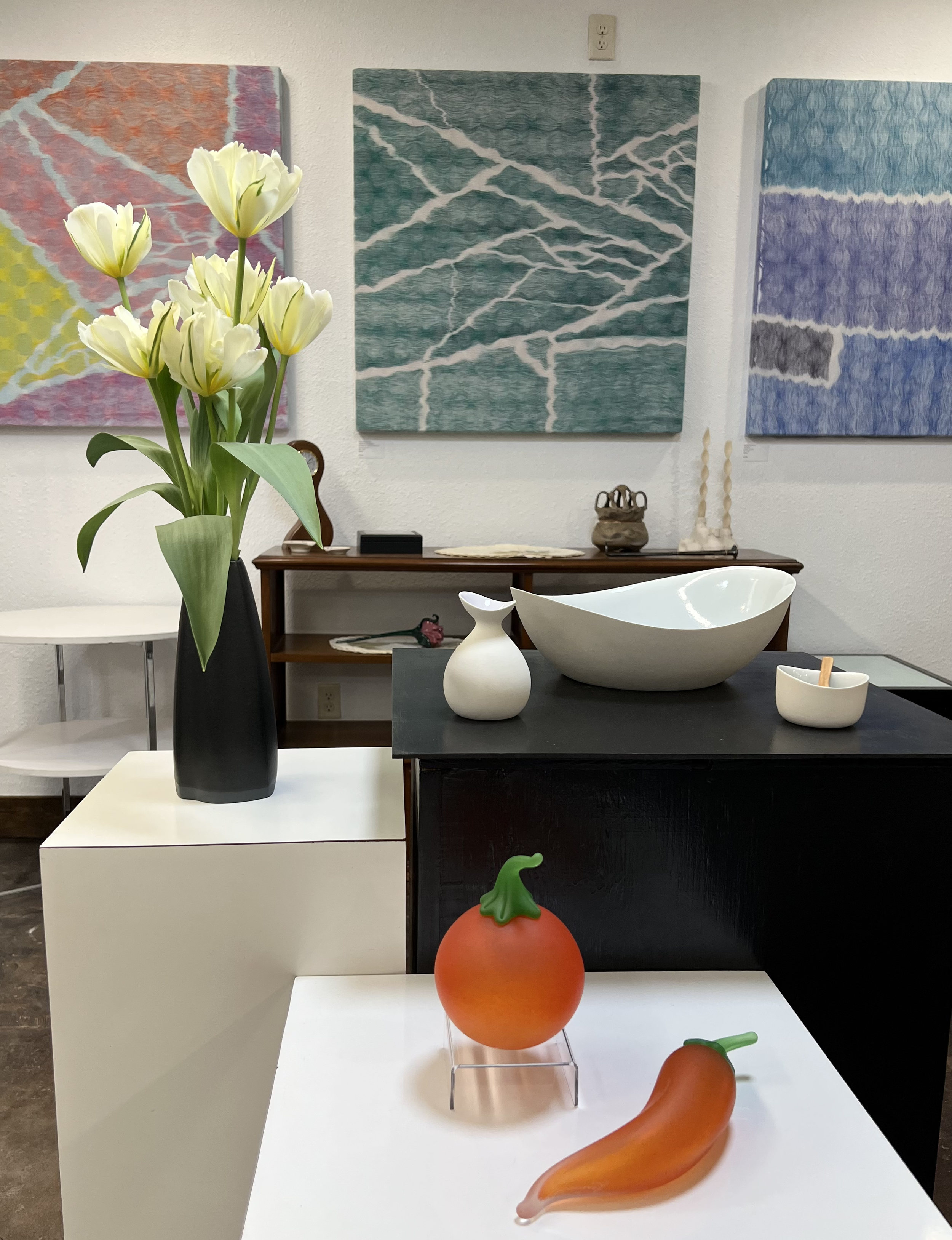 A still life display of art and decor, including a black vase with yellow tulips, white ceramic vases, a large white bowl, and the orange vegetable shaped objects with green tops on white and black tables. In the background, three colorful abstract paintings are hung on a white wall.