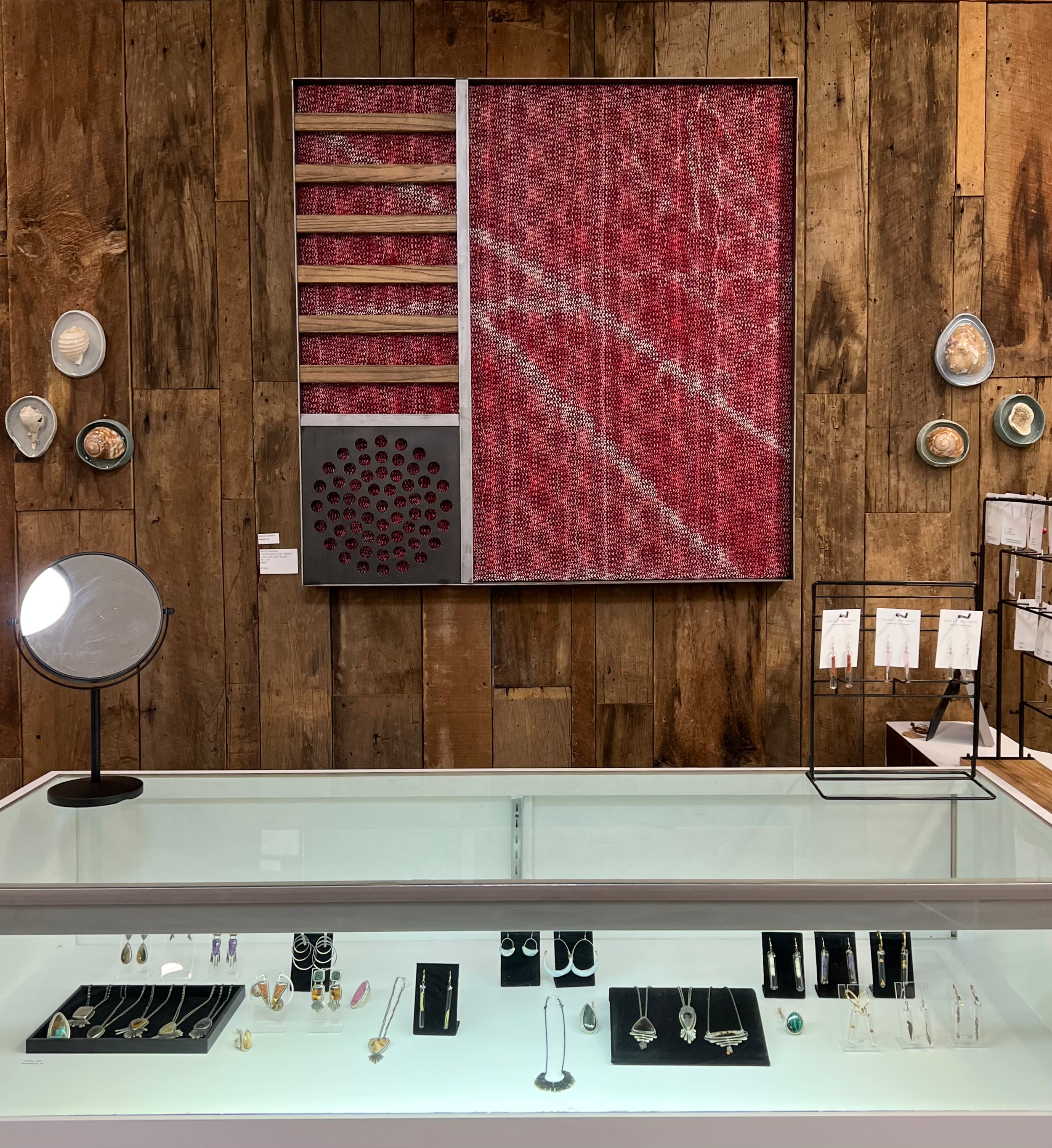 Display of jewelry including rings, earrings, necklaces, and bracelets in a glass case on a white surface with a wooden wall background featuring a steel framed weaving and other wall art.