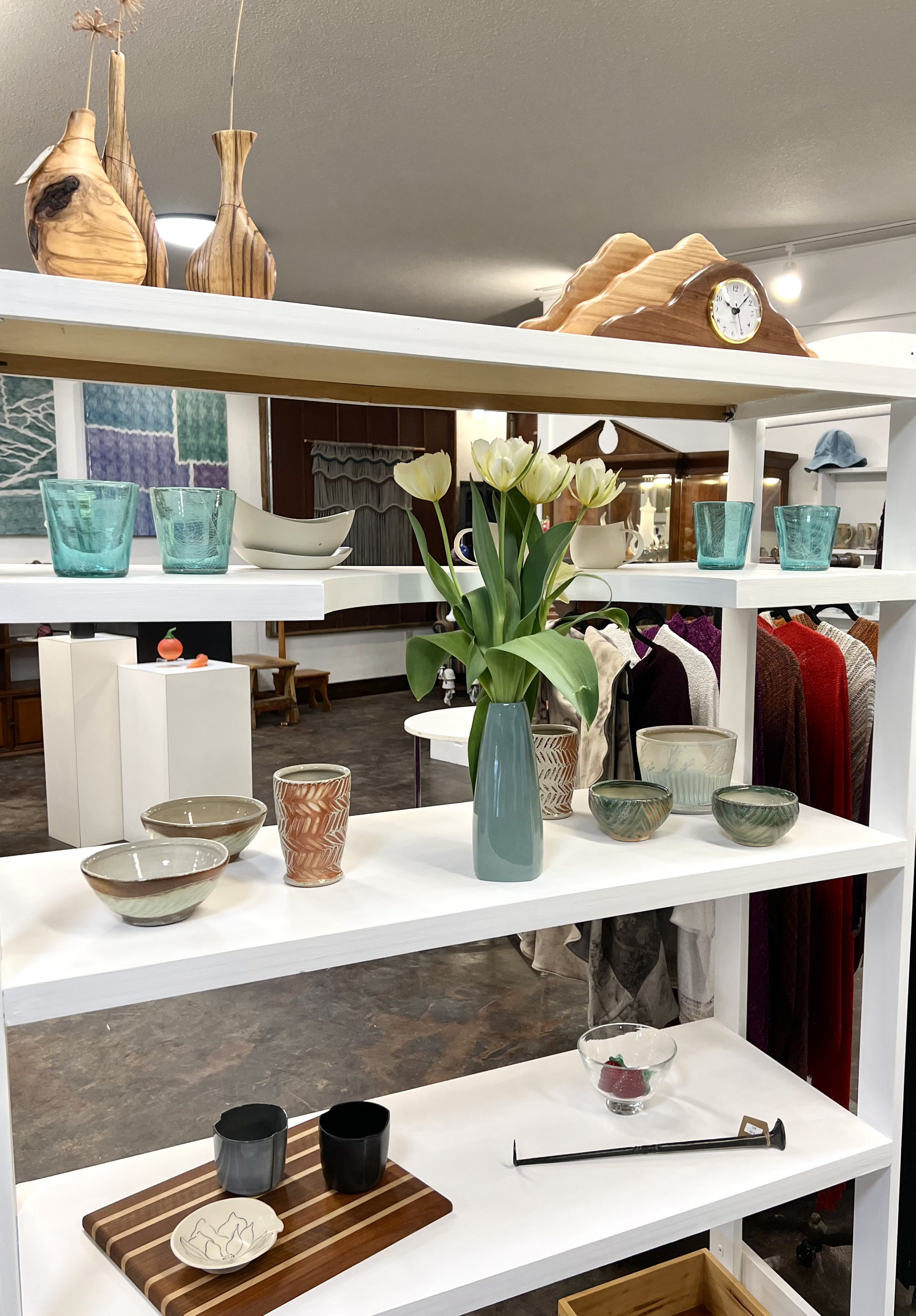 Display of various decorative items including bowls, cups, mugs, and vases on white shelves in a store.