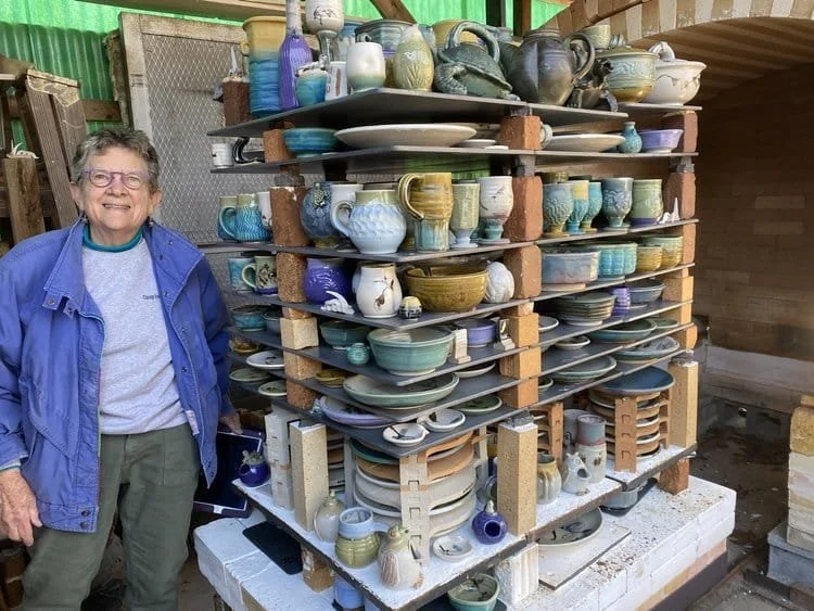 Ceramic artist Cynthia Bringle standing next to a large outdoor kiln filled with ceramic pottery and cups arranged on multiple shelves.