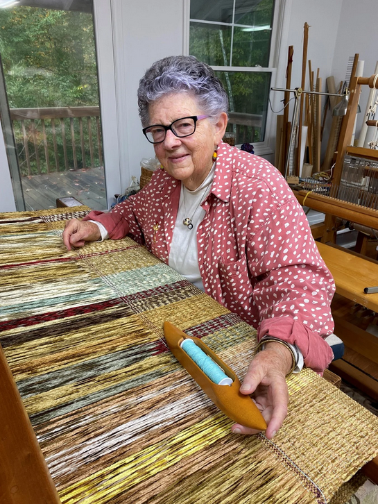 Textile artist Edwina Bringle sitting at a wooden table with a colorful woven textile in front of her, holding a brown wooden tool with blue thread, inside a room with weaving looms in the background.