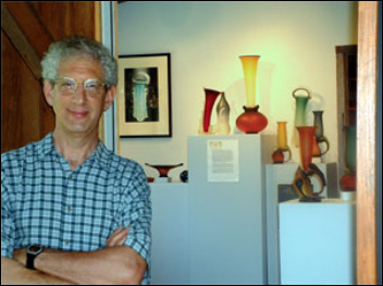 Glass artist Rob Robert Levin in an art gallery next to display cases with colorful glass vases and sculptures.