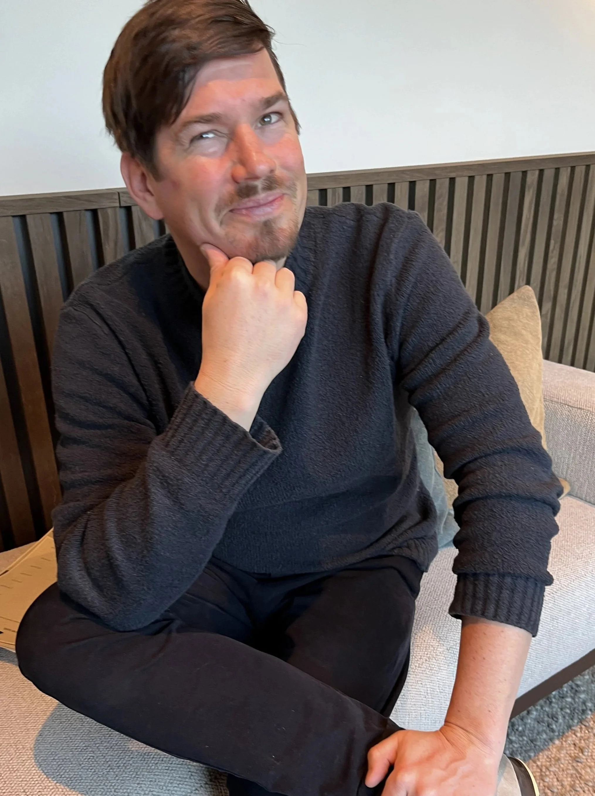 Ein lachender Mann mit dunklem Haar und Bart sitzt auf einer Couch in einem modernen Raum, trägt eine schwarze Strickjacke und schwarze Hosen, macht eine nachdenkliche Pose mit der Hand am Kinn.